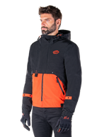 Uomo che indossa il parka impermeabile Alpinestars Mootant in Spicy Orange e nero, una giacca da moto modulare con strato superiore nero con cappuccio e sezione inferiore arancione brillante, abbinata a guanti da guida e pantaloni neri.