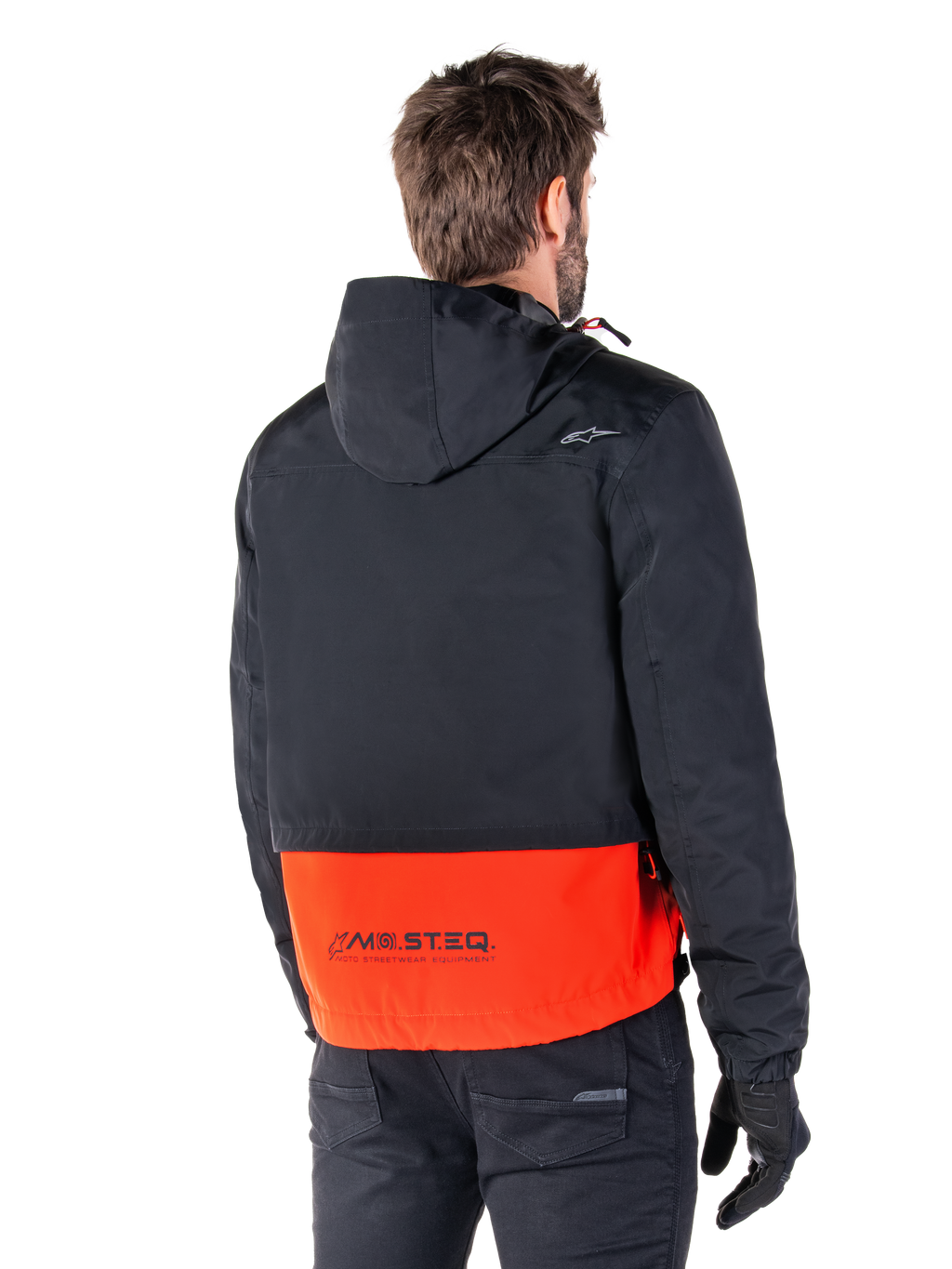 Alpinestars Mootant Waterproof Parka, Arancione Spicy e Nero, vista posteriore su un modello maschile, caratterizzato da una parte superiore nera con cappuccio e una sezione inferiore arancione vivace con marchio MO.ST.EQ., giacca da moto urban contemporary indossata con pantaloni e guanti neri
