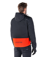 Alpinestars Mootant Waterproof Parka, Arancione Spicy e Nero, vista posteriore su un modello maschile, caratterizzato da una parte superiore nera con cappuccio e una sezione inferiore arancione vivace con marchio MO.ST.EQ., giacca da moto urban contemporary indossata con pantaloni e guanti neri