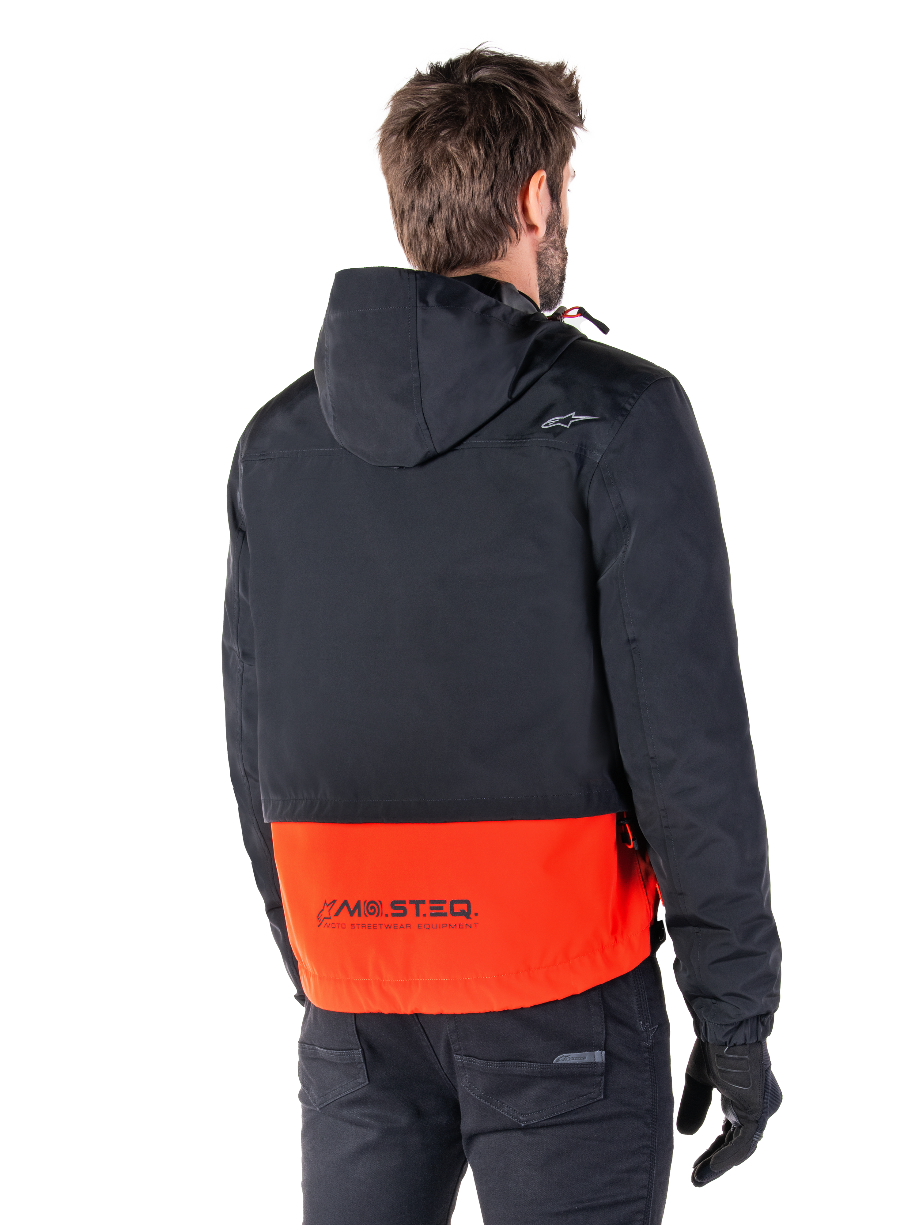 Alpinestars Mootant Waterproof Parka, Arancione Spicy e Nero, vista posteriore su un modello maschile, caratterizzato da una parte superiore nera con cappuccio e una sezione inferiore arancione vivace con marchio MO.ST.EQ., giacca da moto urban contemporary indossata con pantaloni e guanti neri