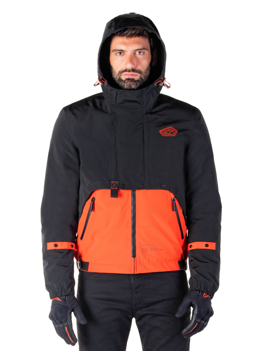 Alpinestars Mootant Waterproof Parka, giacca da moto, arancione e nero, design modulare per tutte le stagioni con cappuccio, tasche anteriori con zip e dettagli con cinturini arancioni sulle maniche, indossata da un modello con guanti e pantaloni neri