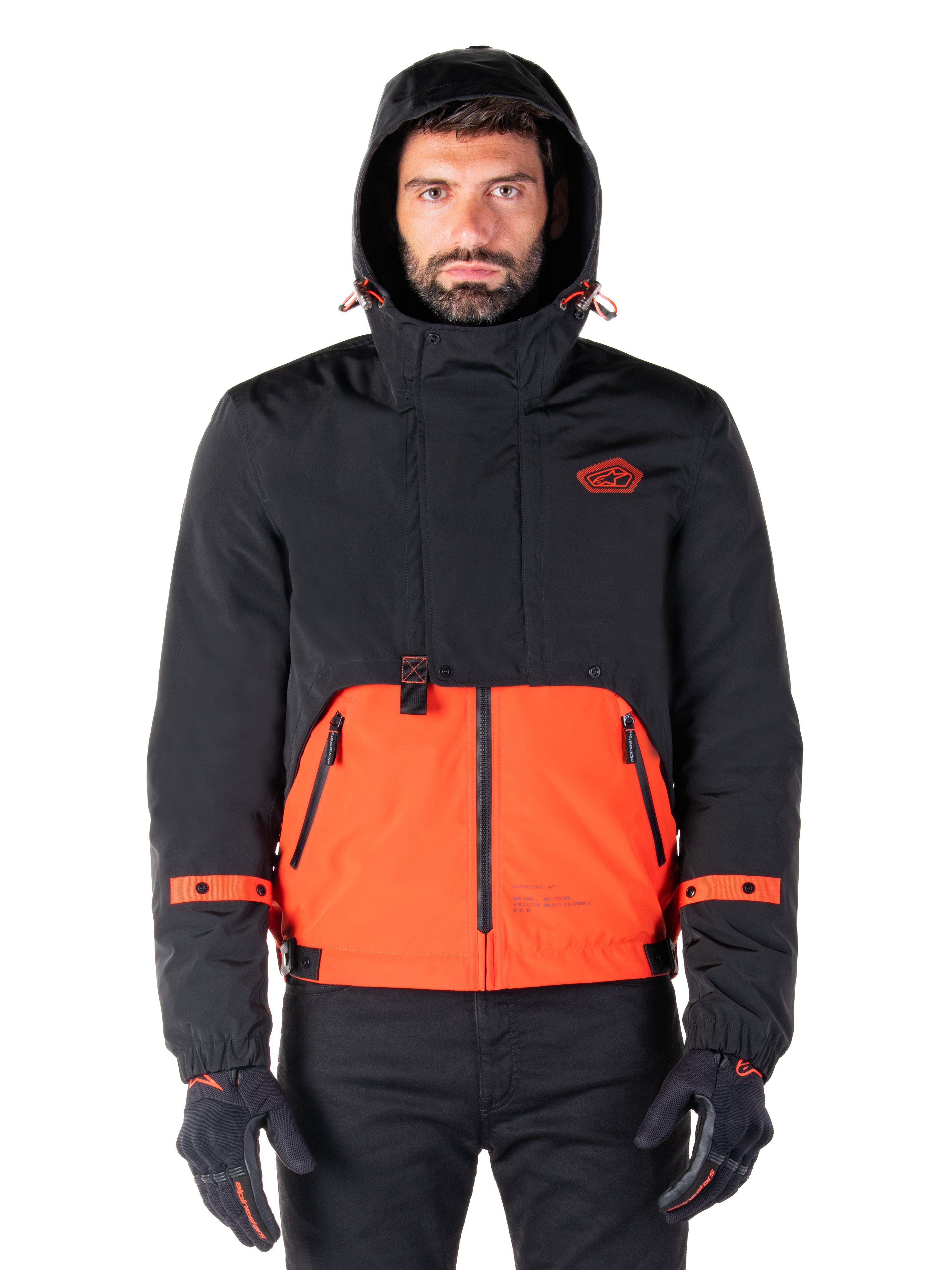 Alpinestars Mootant Waterproof Parka, giacca da moto, arancione e nero, design modulare per tutte le stagioni con cappuccio, tasche anteriori con zip e dettagli con cinturini arancioni sulle maniche, indossata da un modello con guanti e pantaloni neri