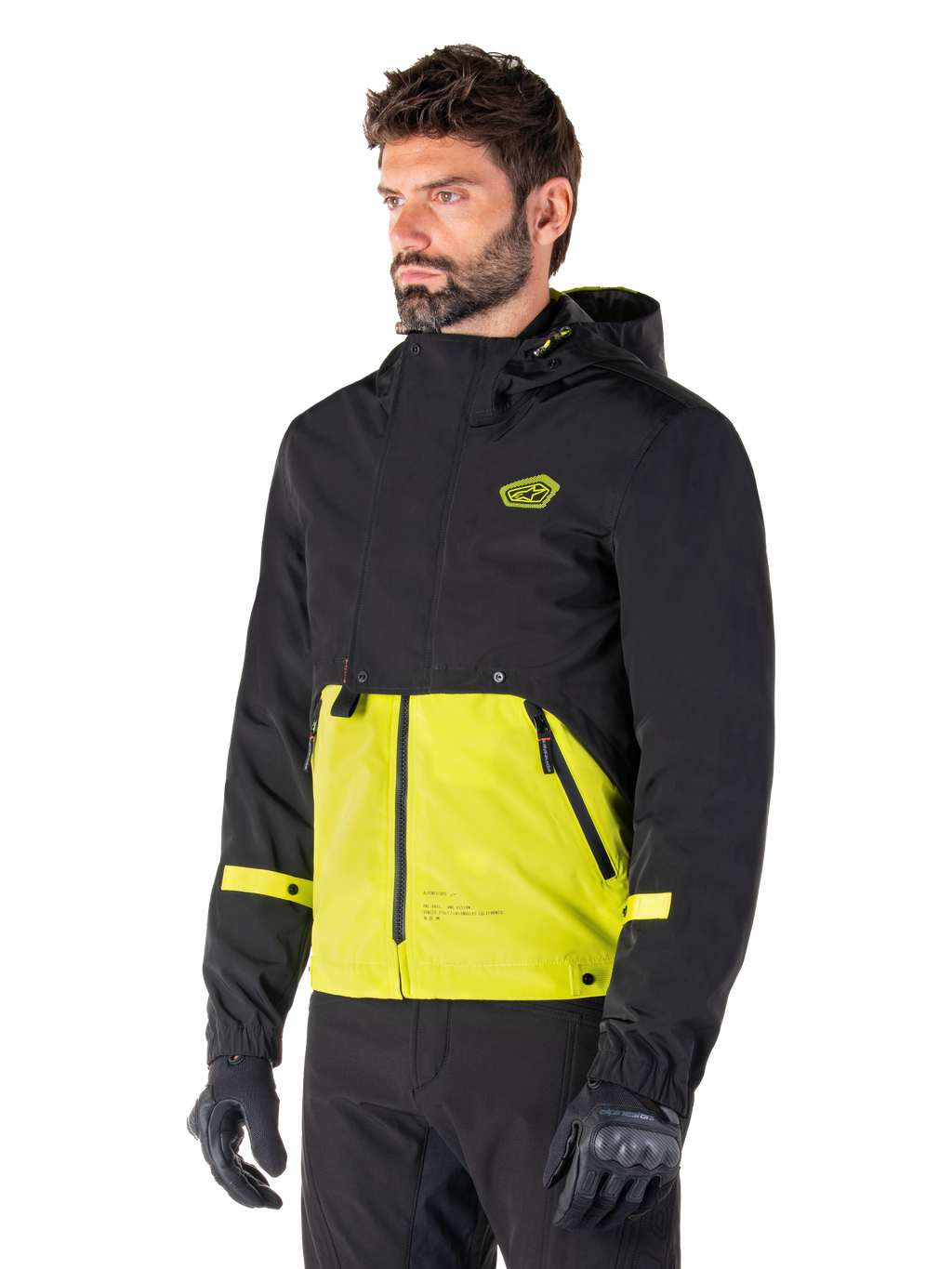 Alpinestars Mootant Waterproof Parka in Lime Punch Black, giacca da moto modulare indossata da un uomo con guanti e pantaloni da guida neri, caratterizzata da una sezione superiore e maniche nere con un pannello inferiore verde lime vivace, design con cappuccio dall'estetica utilitaristica di ispirazione militare.