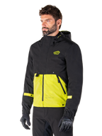 Alpinestars Mootant Waterproof Parka in Lime Punch Black, giacca da moto modulare indossata da un uomo con guanti e pantaloni da guida neri, caratterizzata da una sezione superiore e maniche nere con un pannello inferiore verde lime vivace, design con cappuccio dall'estetica utilitaristica di ispirazione militare.