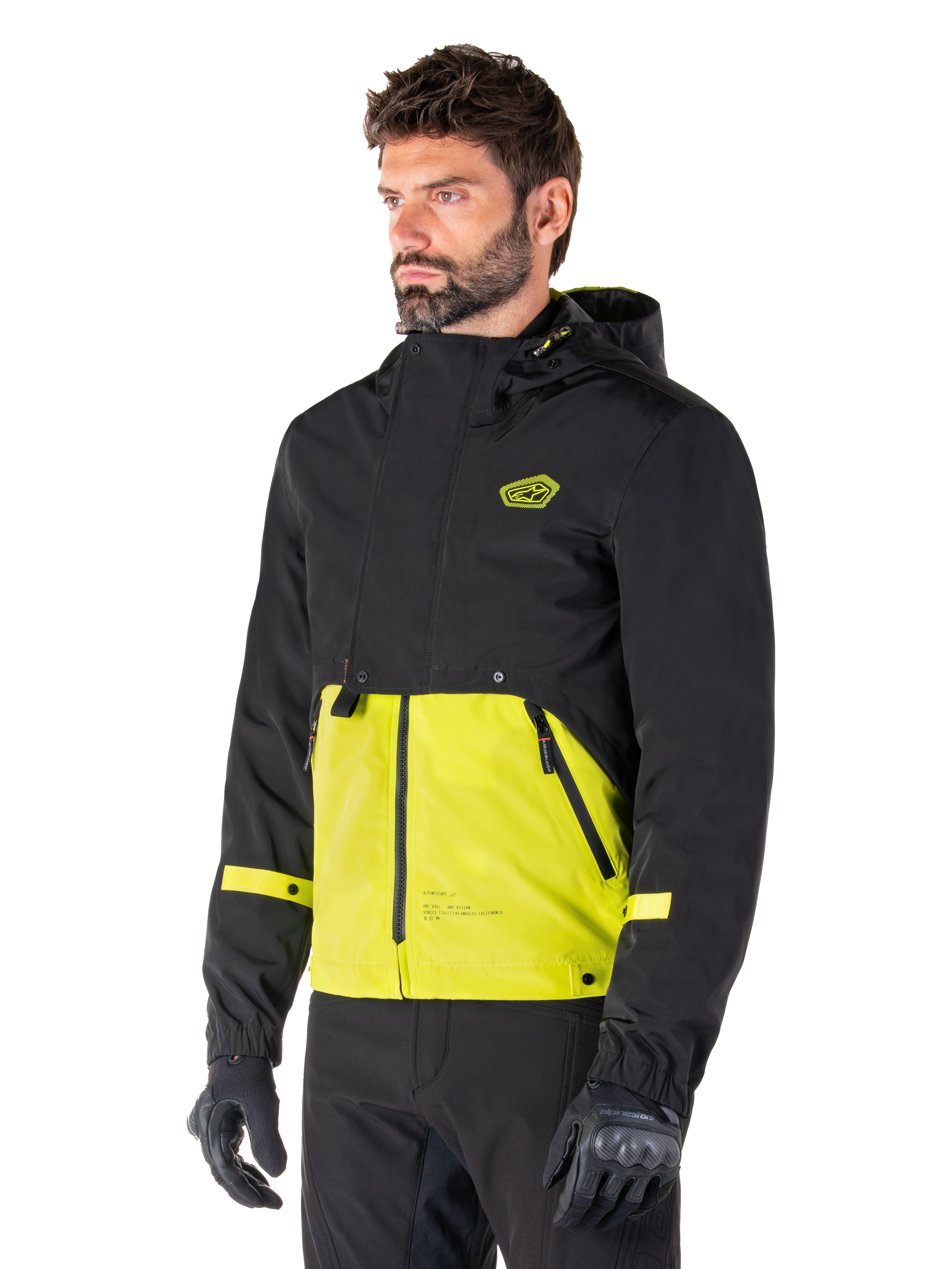 Alpinestars Mootant Waterproof Parka in Lime Punch Black, giacca da moto modulare indossata da un uomo con guanti e pantaloni da guida neri, caratterizzata da una sezione superiore e maniche nere con un pannello inferiore verde lime vivace, design con cappuccio dall'estetica utilitaristica di ispirazione militare.