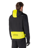 Alpinestars Mootant Waterproof Parka, vista posteriore, Lime Punch e nero, caratterizzato da una parte superiore nera con pannello del cappuccio e parte inferiore della schiena giallo lime, marchio MO.ST.EQ. sull'orlo, indossato con pantaloni da moto e guanti neri, giacca da moto urban contemporary