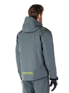 Alpinestars Morush Waterproof Parka in Urban Gray, vista posteriore, con marchio MO.ST.EQ. giallo sulla parte bassa della schiena e sulla spalla, design minimalista con cappuccio, indossato con pantaloni da moto grigi coordinati e guanti neri, abbigliamento urban streetwear da moto.