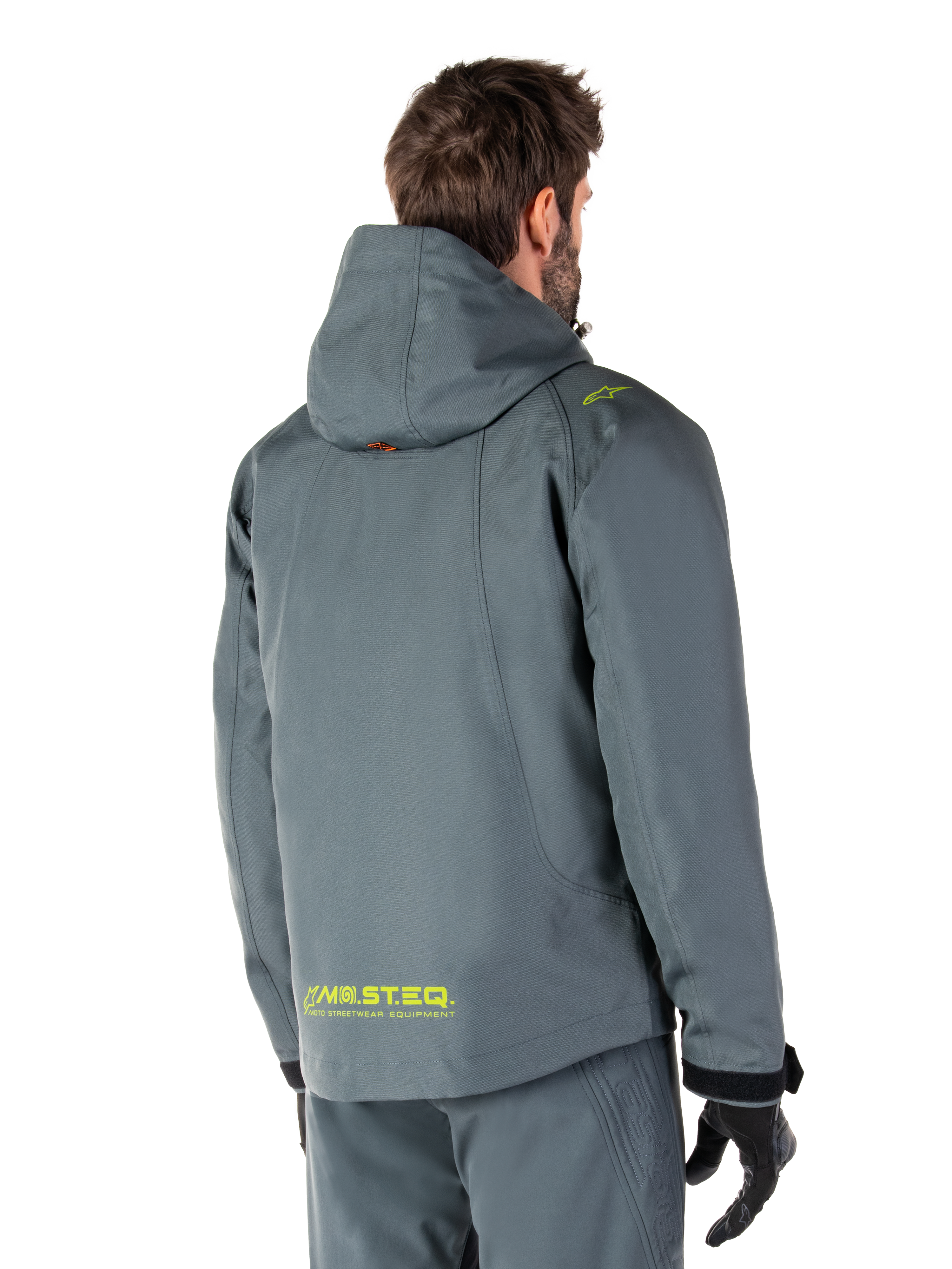 Alpinestars Morush Waterproof Parka in Urban Gray, vista posteriore, con marchio MO.ST.EQ. giallo sulla parte bassa della schiena e sulla spalla, design minimalista con cappuccio, indossato con pantaloni da moto grigi coordinati e guanti neri, abbigliamento urban streetwear da moto.