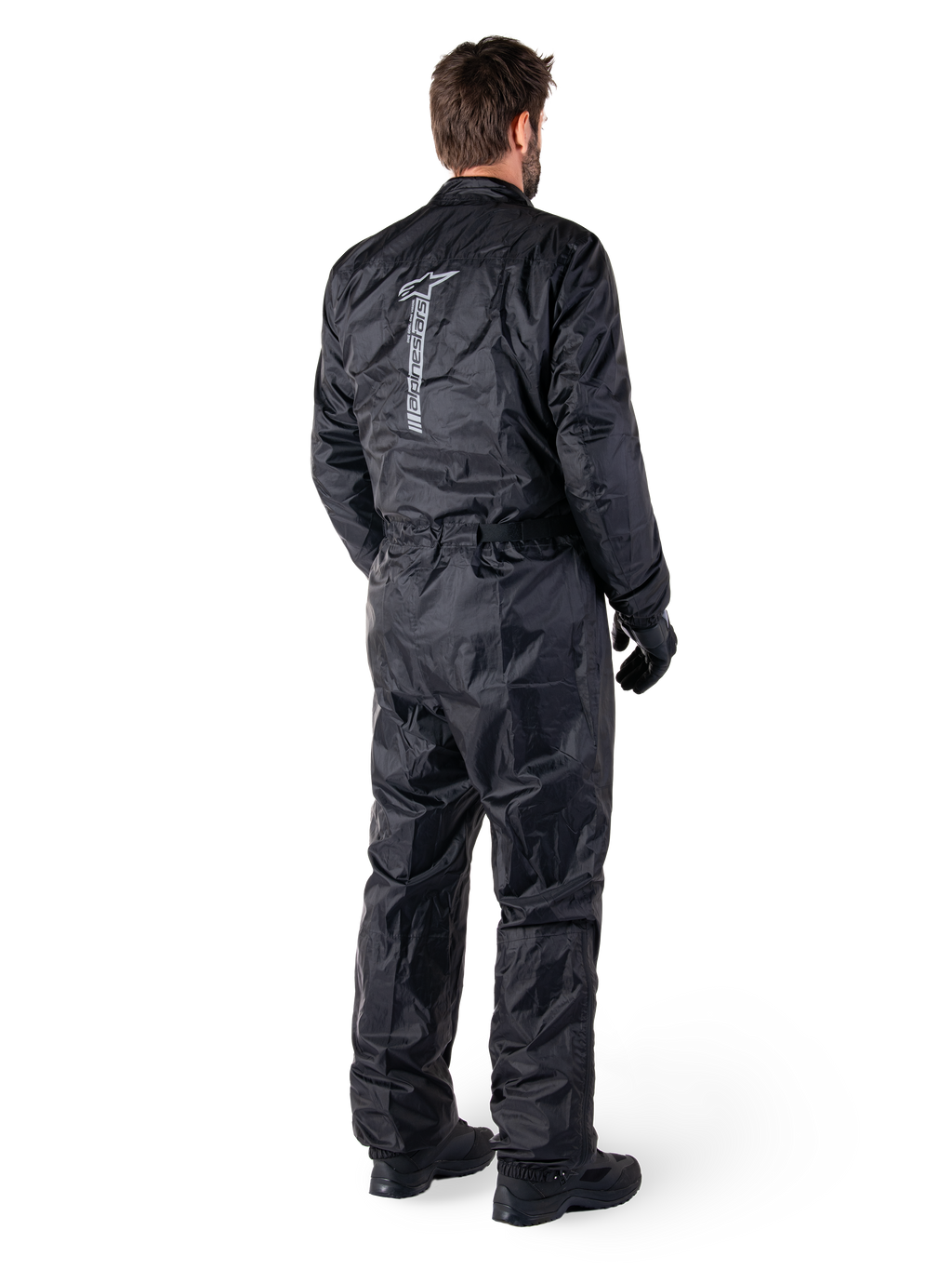 Tuta Alpinestars Hurricane Rain V2, tuta da moto antipioggia monopezzo impermeabile, nera, vista posteriore con grande logo riflettente verticale, tessuto leggero e resistente al vento, indossata con guanti e stivali neri