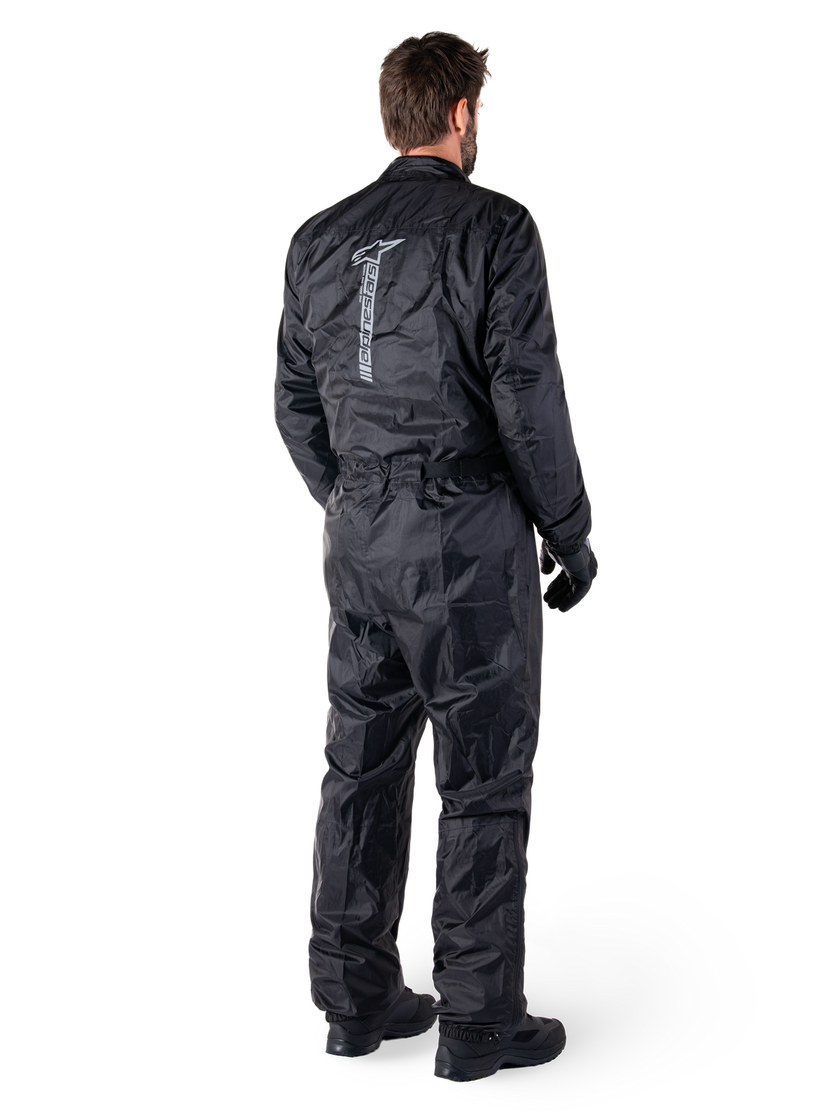 Tuta Alpinestars Hurricane Rain V2, tuta da moto antipioggia monopezzo impermeabile, nera, vista posteriore con grande logo riflettente verticale, tessuto leggero e resistente al vento, indossata con guanti e stivali neri