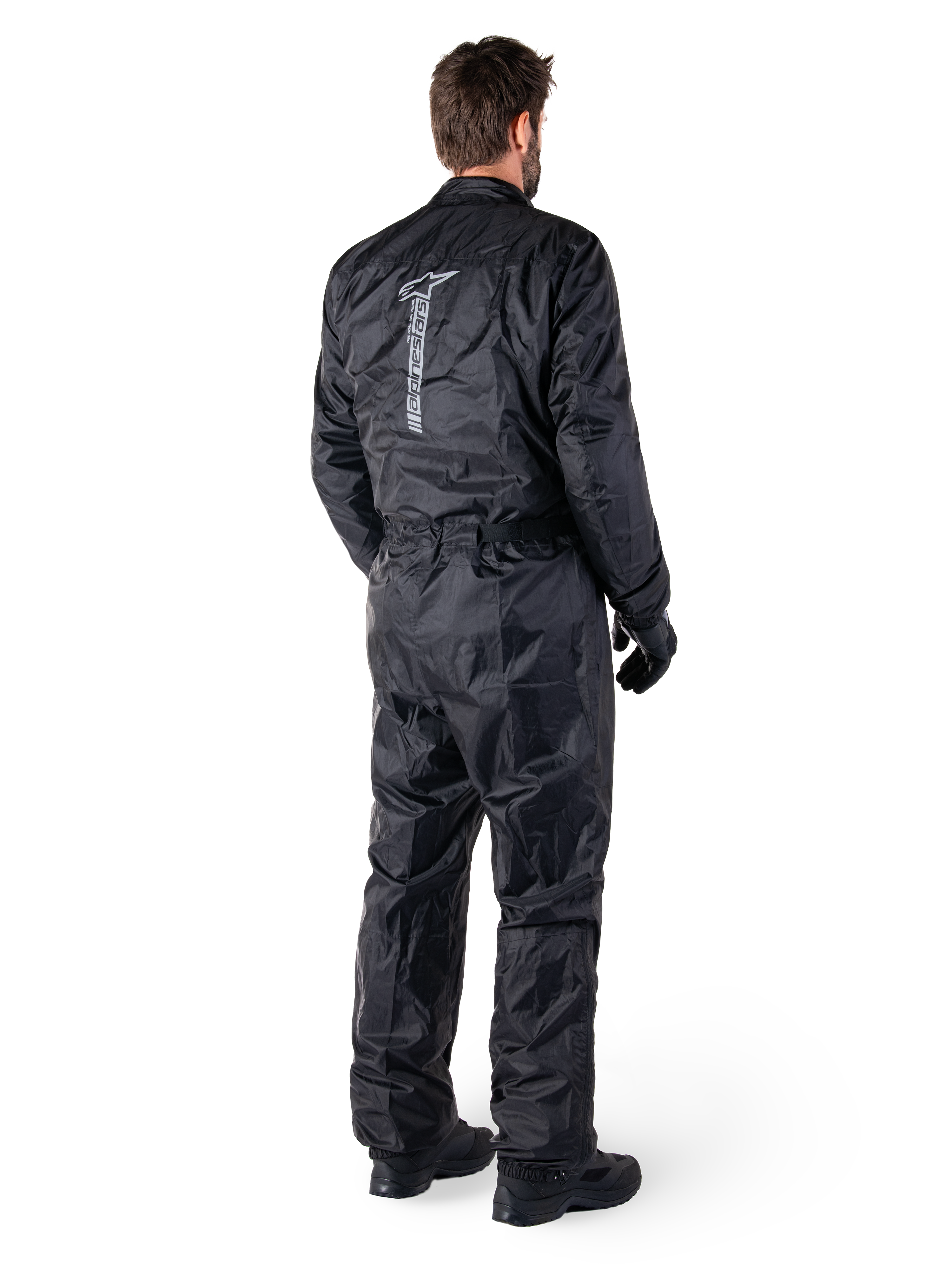 Tuta Alpinestars Hurricane Rain V2, tuta da moto antipioggia monopezzo impermeabile, nera, vista posteriore con grande logo riflettente verticale, tessuto leggero e resistente al vento, indossata con guanti e stivali neri