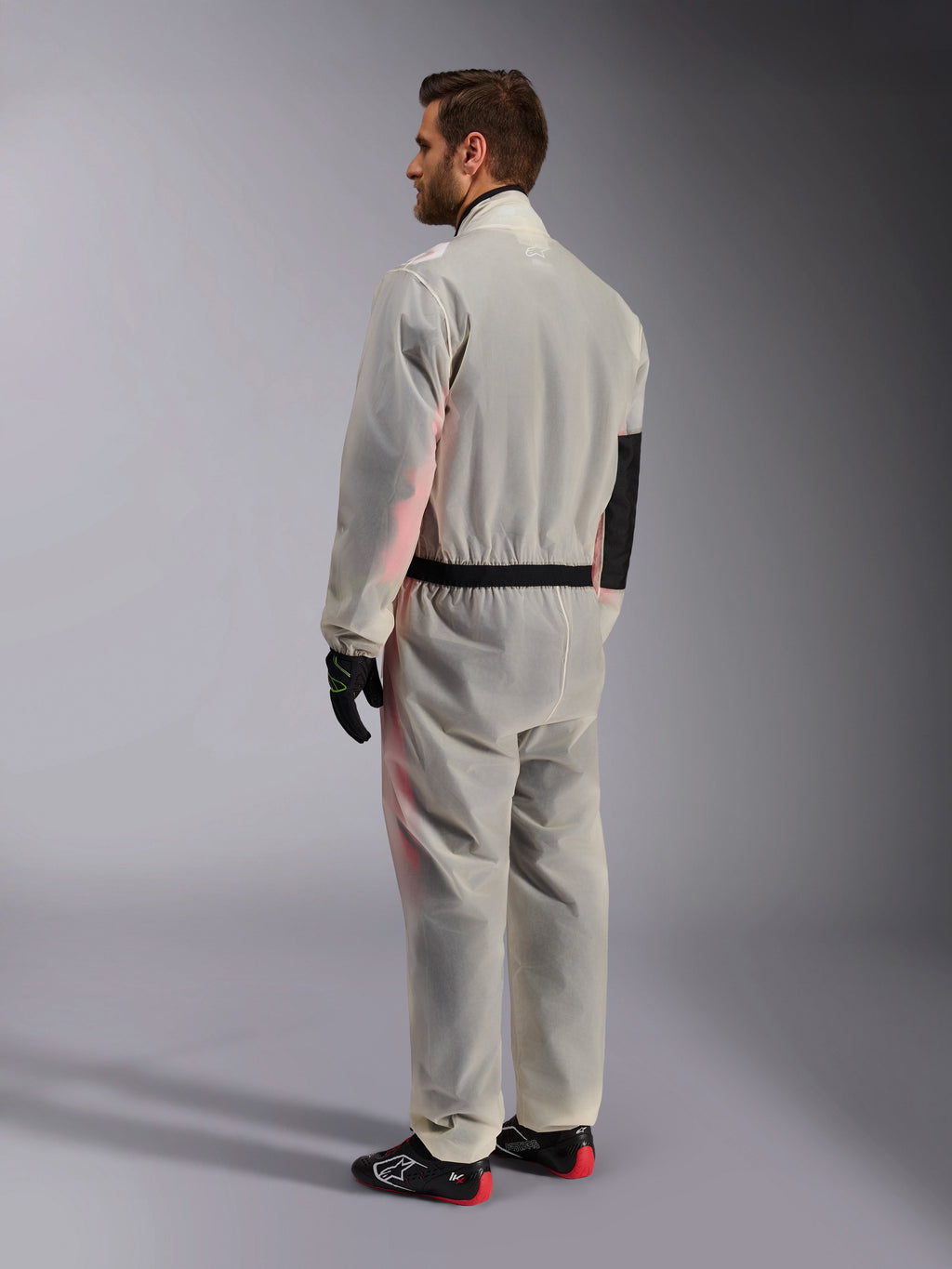Alpinestars Kart Rain Suit, sovratuta trasparente impermeabile da kart, trasparente, vista posteriore di tre quarti, con pannello di rinforzo nero sul gomito e vita elasticizzata, indossata sopra una tuta da gara con scarpe nere e rosse, progettata per il karting su bagnato