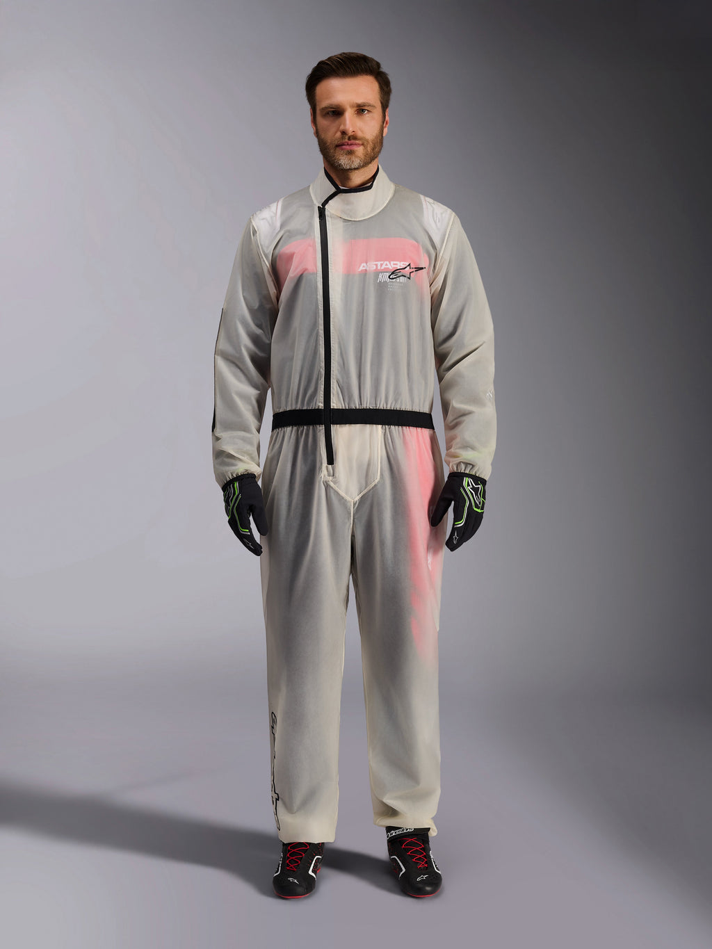 Alpinestars Kart Rain Suit, trasparente, sovratuta impermeabile trasparente indossata da un modello, cerniera asimmetrica nera e logo sul petto, tuta da corsa rossa e bianca visibile sotto, guanti neri e verde neon e stivali da corsa neri