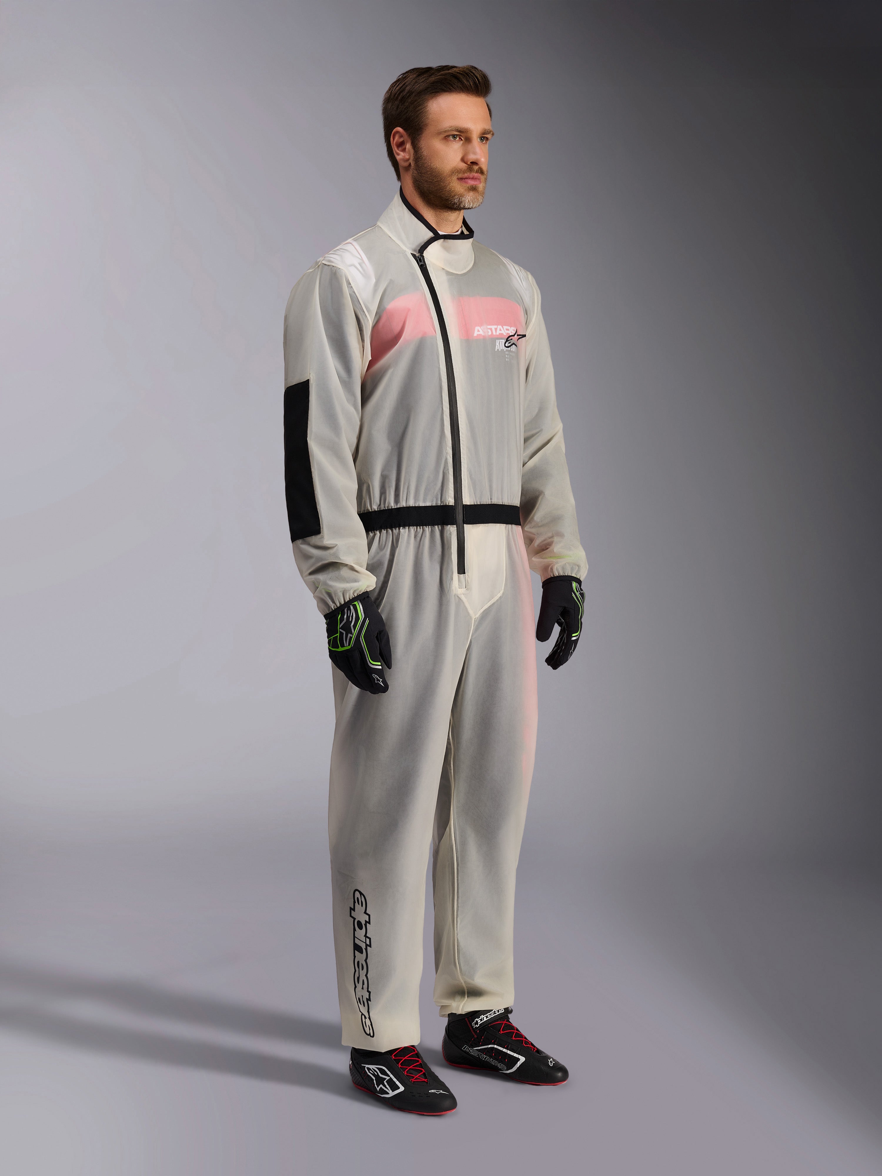Alpinestars Kart Rain Suit, sovratuta da kart impermeabile trasparente, materiale semitrasparente che mostra una tuta da gara rosa e bianca sottostante, con cerniera asimmetrica nera, rinforzo sul gomito e fascia nera in vita, indossata con guanti da kart e scarpe da racing neri.