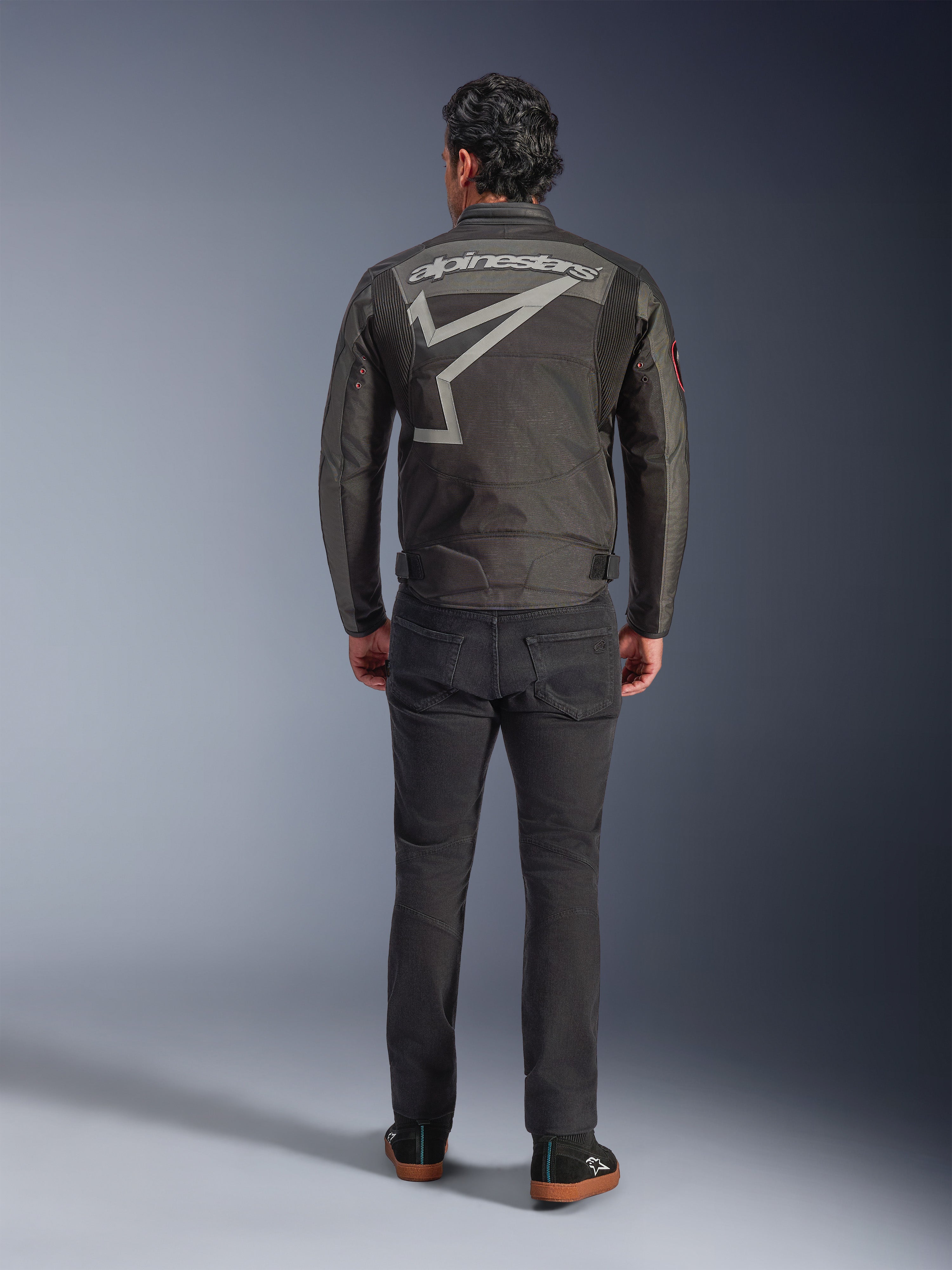 Giacca Alpinestars Axton, giacca da moto, Nero Antracite Rosso Tangerine, vista posteriore con grande logo Alpinestars grigio e grafica a stella, pannelli a fisarmonica sulle spalle, indossata con pantaloni neri e scarpe casual nere.