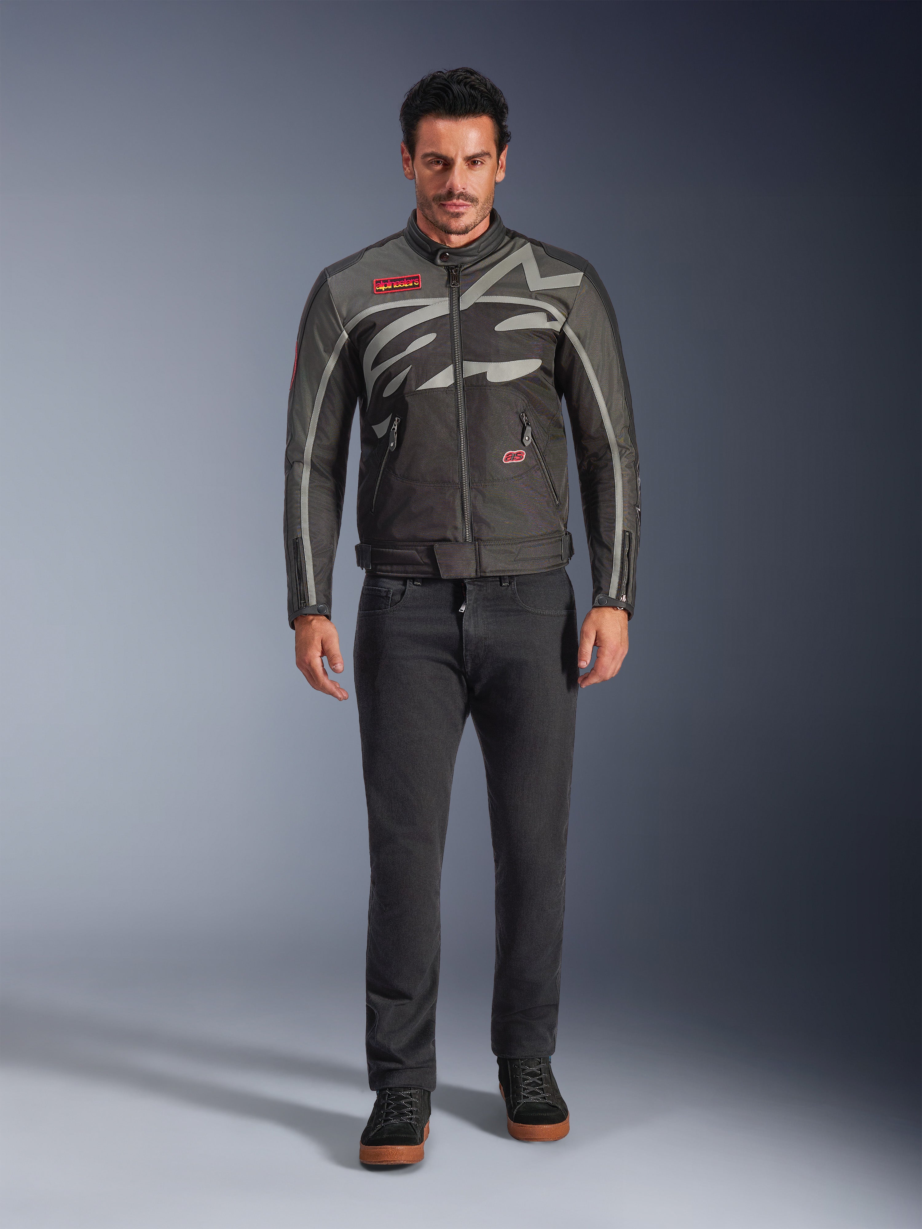 Giacca Alpinestars Axton, giacca da moto, nero antracite rosso tangerine, indossata da un modello con pantaloni scuri, con logo Alpinestars grigio avvolgente, patch del marchio rosse e guscio in tessuto resistente all'acqua per la guida urbana