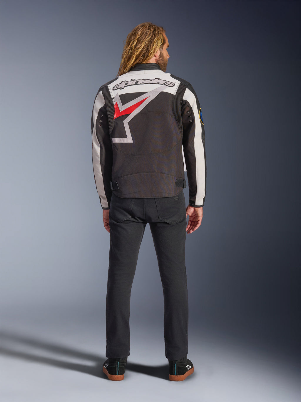 Giacca Alpinestars Axton, giacca da moto, nero, bianco sporco e rosso brillante, vista posteriore su un modello, con un grande logo a stella Alpinestars sul retro, pannelli delle maniche bianco sporco, indossata con pantaloni neri, design ispirato all'abbigliamento racing vintage
