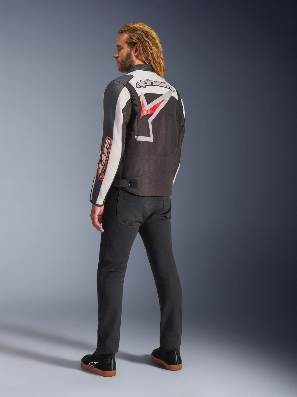Giacca Alpinestars Axton, giacca da moto, nero, bianco sporco e rosso acceso, vista posteriore indossata da un modello, con grande logo a stella Alpinestars e scritta sulla schiena, design d'ispirazione racing vintage con pannelli a fisarmonica, abbinata a pantaloni neri e sneakers