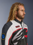 Uomo con capelli lunghi e barba che indossa la Giacca Alpinestars Axton, giacca da moto, nero, bianco sporco e rosso vivace, design ispirato al racing vintage con patch logate e logo avvolgente, vista di profilo ravvicinata, stile contemporaneo urbano