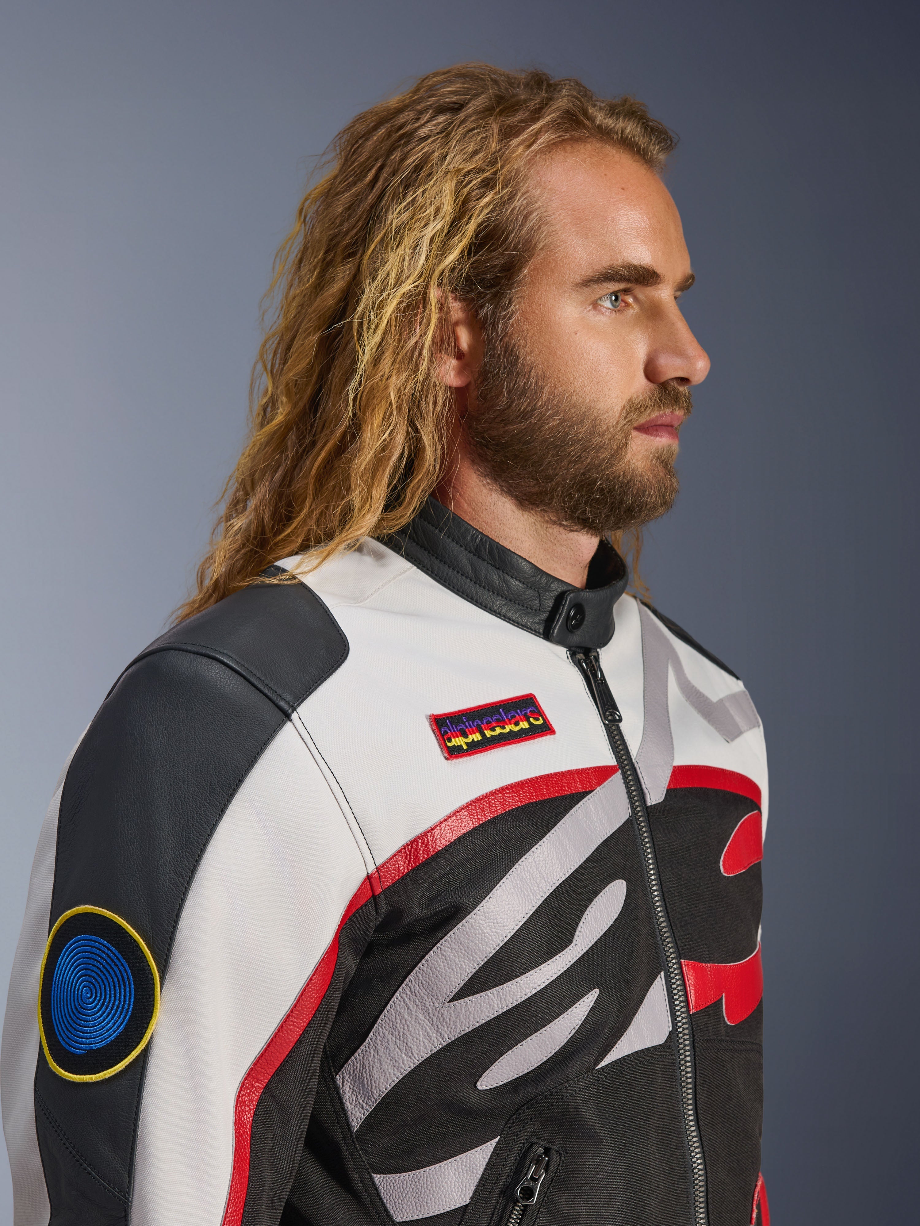 Uomo con capelli lunghi e barba che indossa la Giacca Alpinestars Axton, giacca da moto, nero, bianco sporco e rosso vivace, design ispirato al racing vintage con patch logate e logo avvolgente, vista di profilo ravvicinata, stile contemporaneo urbano