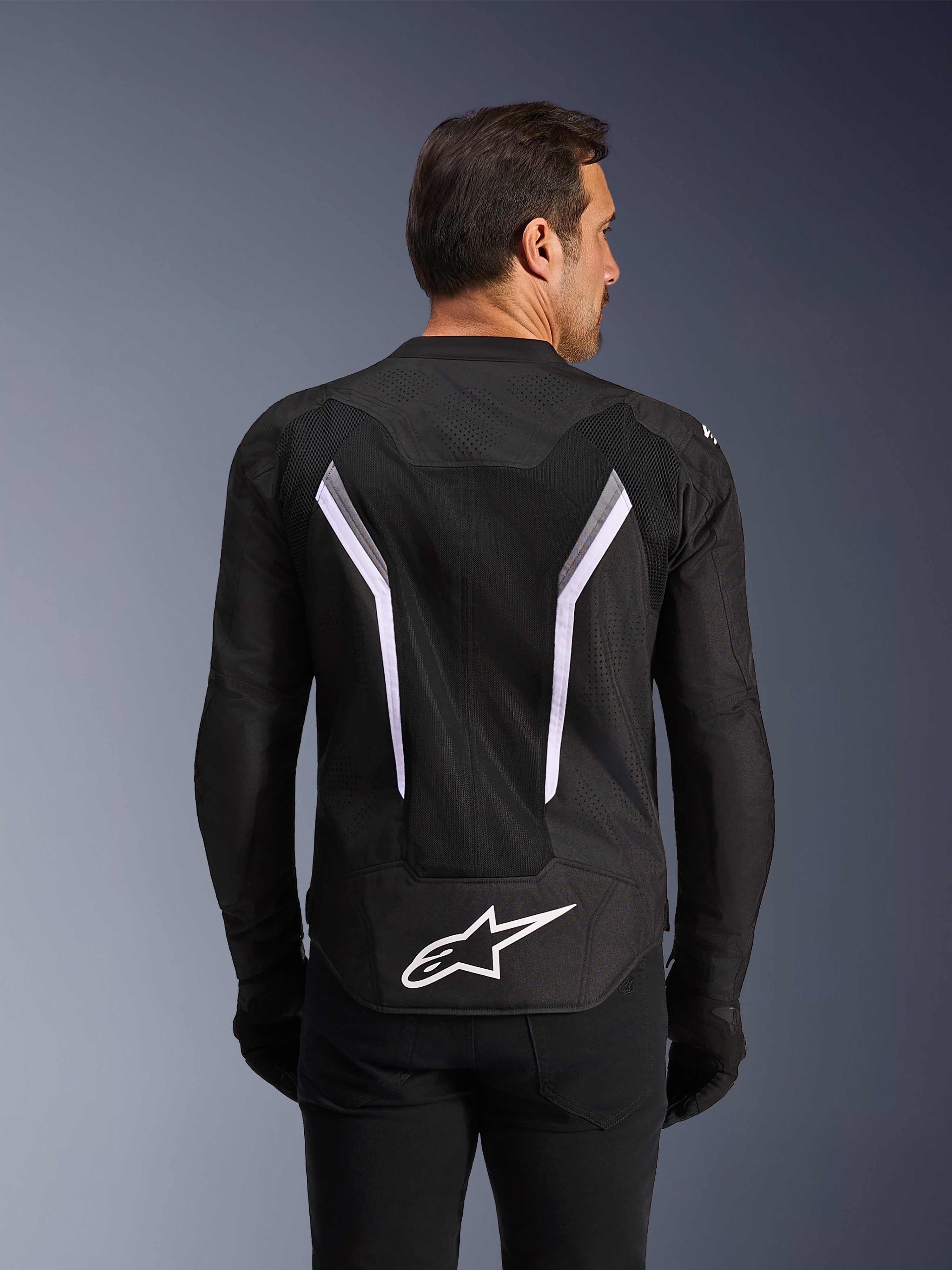 Giacca Alpinestars AST V3 Air, giacca da moto, nera con accenti bianchi e grigio scuro, vista posteriore che mostra ampi pannelli di ventilazione in mesh, grandi strisce bianche a forma di V e logo Alpinestars bianco sulla parte inferiore della schiena, indossata con guanti e pantaloni neri