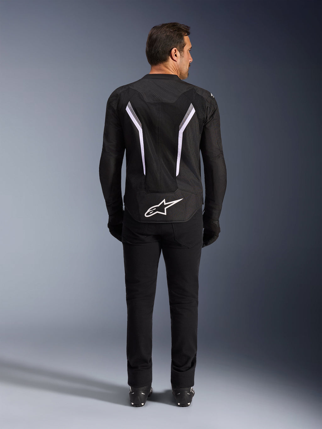 Giacca Alpinestars AST V3 Air, vista posteriore, nero e grigio scuro con accenti bianchi, con grande logo Astar bianco sulla parte inferiore, strisce riflettenti sul retro e costruzione in tessuto mesh, indossata con pantaloni e stivali neri per un look sportivo