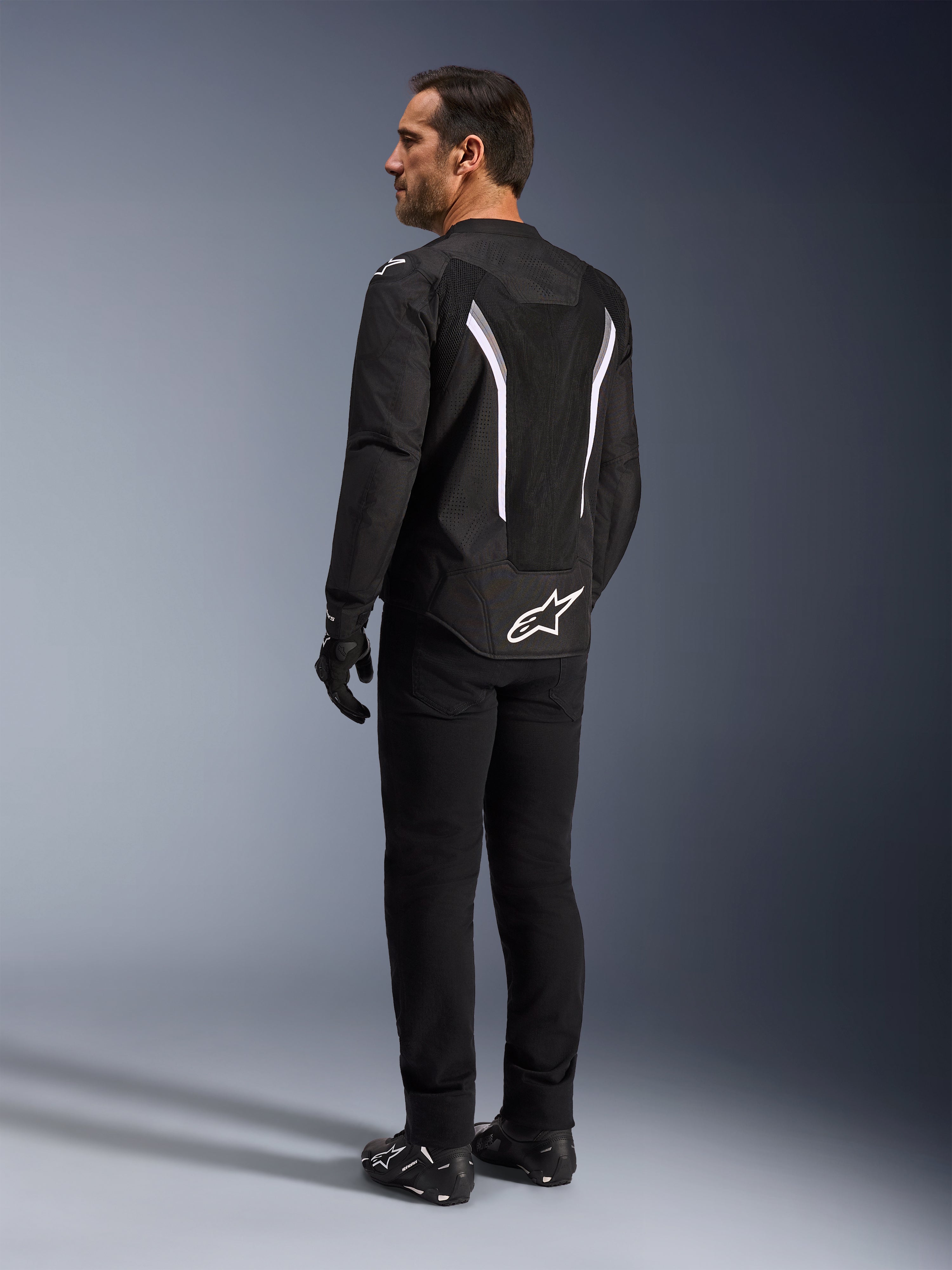 Alpinestars AST V3 Air Jacket, giacca da moto nera e bianca, vista posteriore su modello, con inserti in mesh per il flusso d'aria e logo bianco sulla parte inferiore della schiena, indossata con abbigliamento tecnico da guida nero