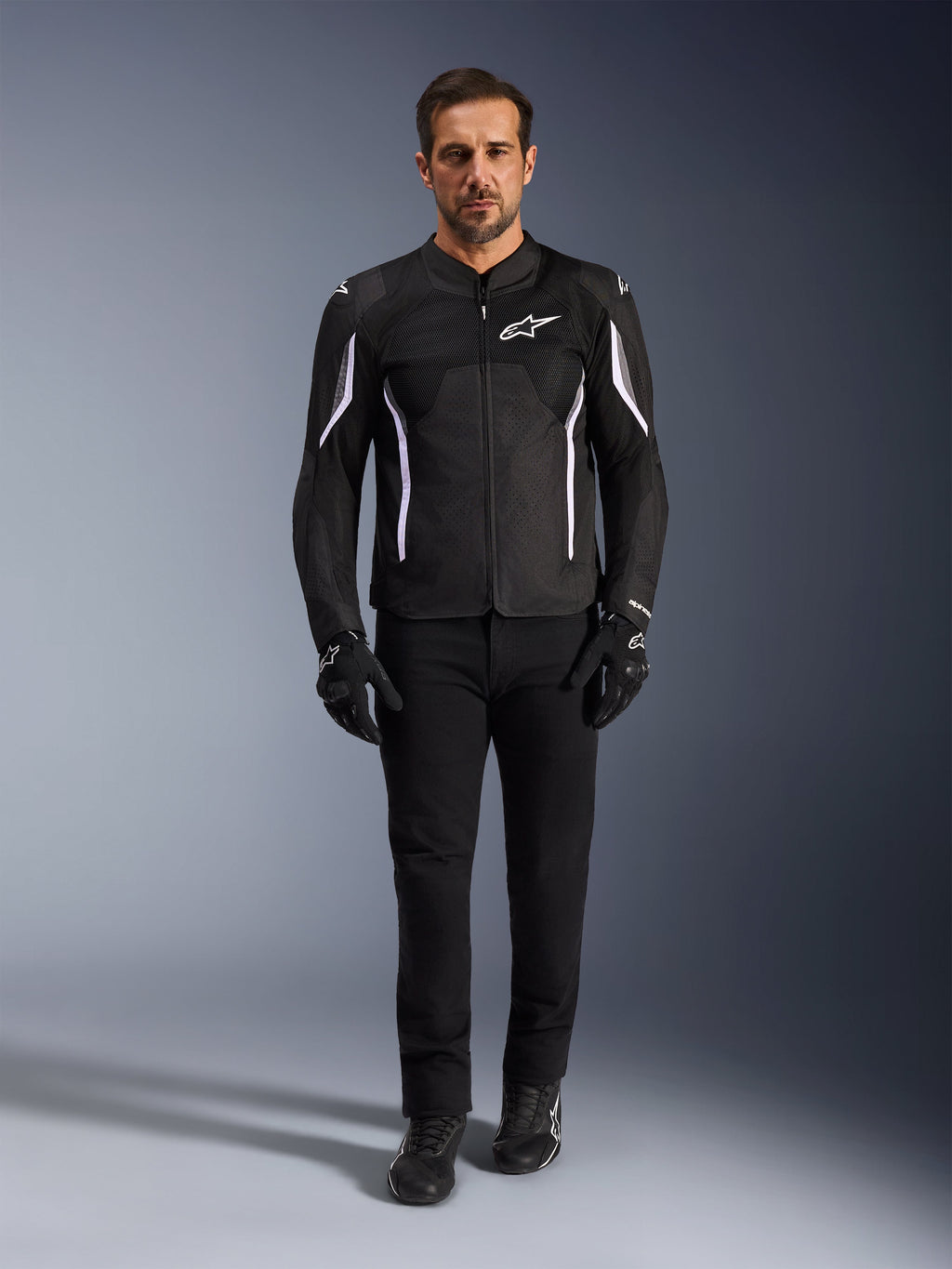 Modello con giacca Alpinestars AST V3 Air in nero con accenti bianchi, con pannelli di ventilazione in mesh e vestibilità sportiva, abbinata a pantaloni tecnici da moto neri, guanti e stivali, vista frontale, abbigliamento tecnico per la guida estiva