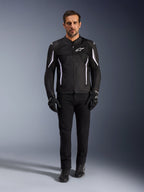 Modello con giacca Alpinestars AST V3 Air in nero con accenti bianchi, con pannelli di ventilazione in mesh e vestibilità sportiva, abbinata a pantaloni tecnici da moto neri, guanti e stivali, vista frontale, abbigliamento tecnico per la guida estiva