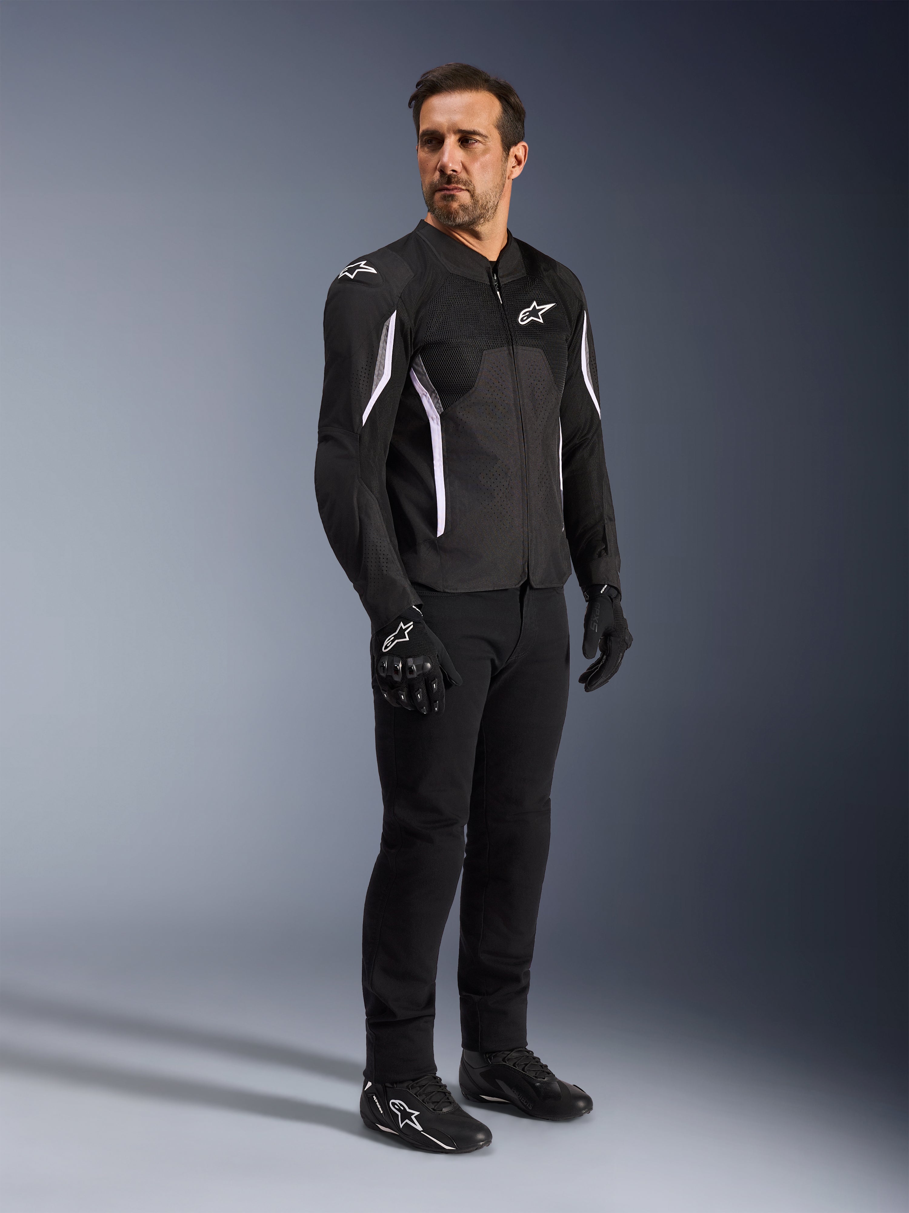 Alpinestars AST V3 Air Jacket, nera con accenti e logo bianchi, giacca da moto leggera con ventilazione in mesh, indossata da un uomo con pantaloni da guida, guanti e scarpe neri, abbigliamento tecnico estivo per guida sportiva e urbana.