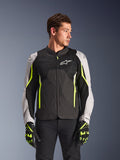 Alpinestars AST V3 Air Jacket, giacca da moto in tessuto, nero e argento con accenti giallo fluo, dotata di ampi pannelli di ventilazione in rete e perforazioni laser, indossata da un modello con guanti coordinati, abbigliamento tecnico sportivo per motociclismo.