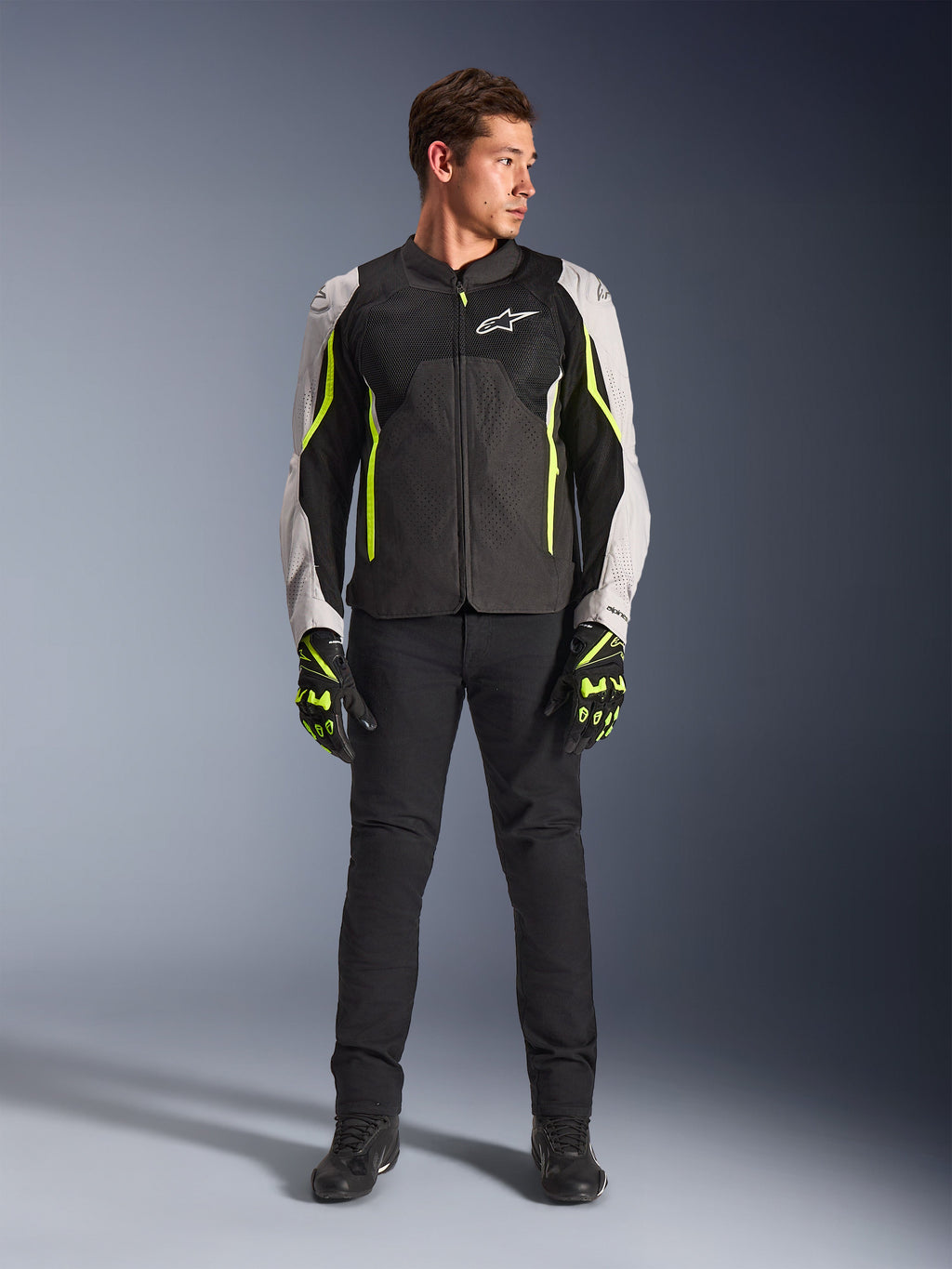 Modello uomo che indossa la giacca Alpinestars AST V3 Air in nero, argento e giallo fluo, con pannelli di ventilazione in rete e accenti fluorescenti, abbinata a pantaloni da moto neri e guanti da moto coordinati, abbigliamento tecnico per guida sportiva.