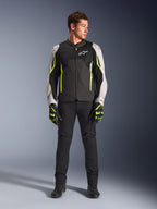 Modello uomo che indossa la giacca Alpinestars AST V3 Air in nero, argento e giallo fluo, con pannelli di ventilazione in rete e accenti fluorescenti, abbinata a pantaloni da moto neri e guanti da moto coordinati, abbigliamento tecnico per guida sportiva.