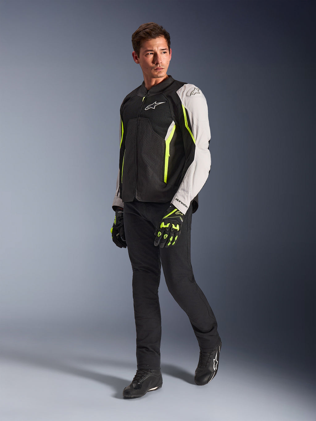 Giacca Alpinestars AST V3 Air, giacca da moto leggera in tessuto nero, argento e giallo fluo, indossata da un modello con pantaloni neri e guanti abbinati, con pannelli in rete e perforazioni laser per la guida sportiva in climi caldi.