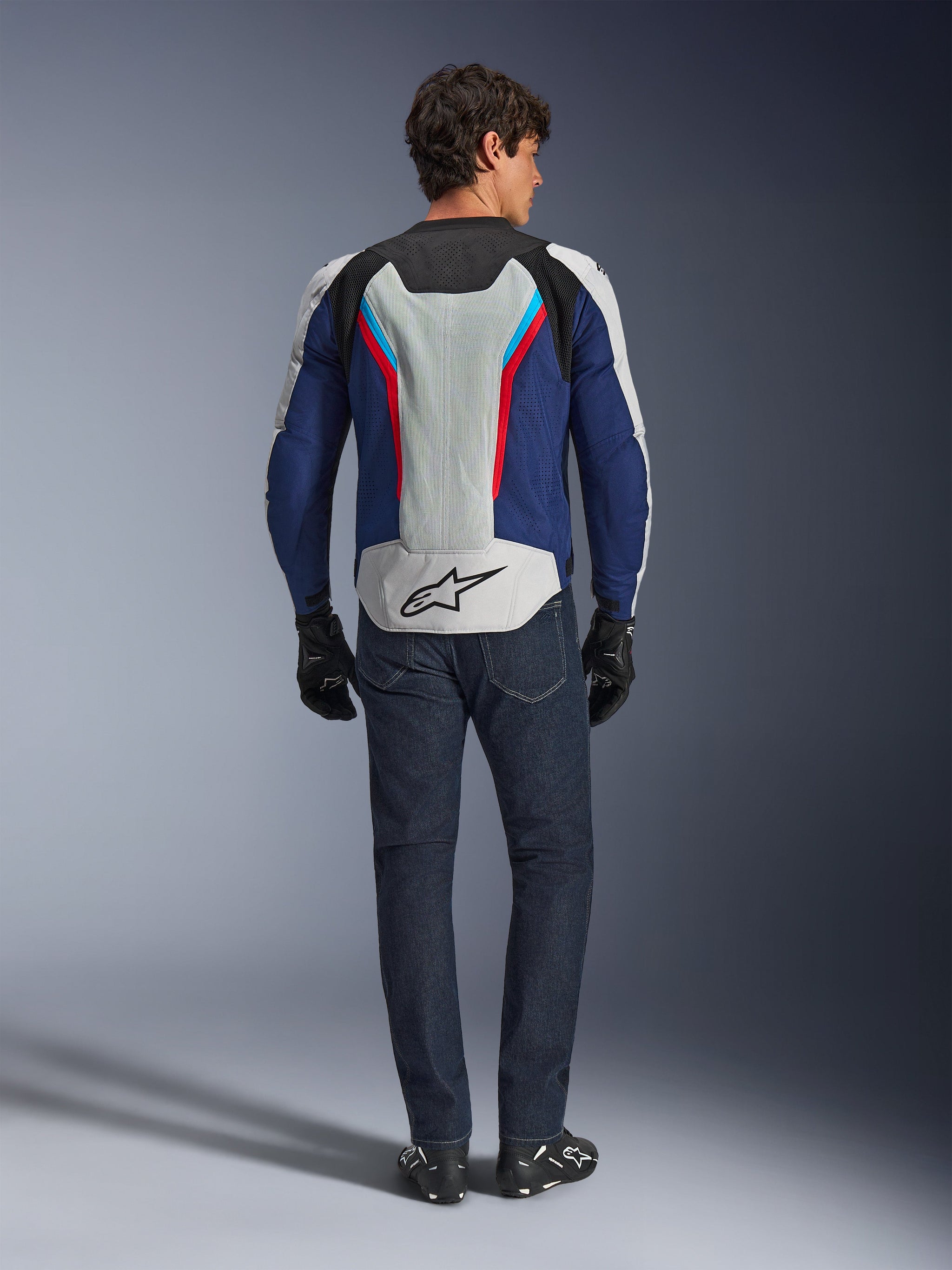 Giacca Alpinestars AST V3 Air, vista posteriore, Silver Deep Blue e Bright Red, con un ampio pannello di ventilazione in rete sulla schiena con logo Alpinestars, indossata con jeans blu scuro e guanti neri, abbigliamento moto sportivo per climi caldi