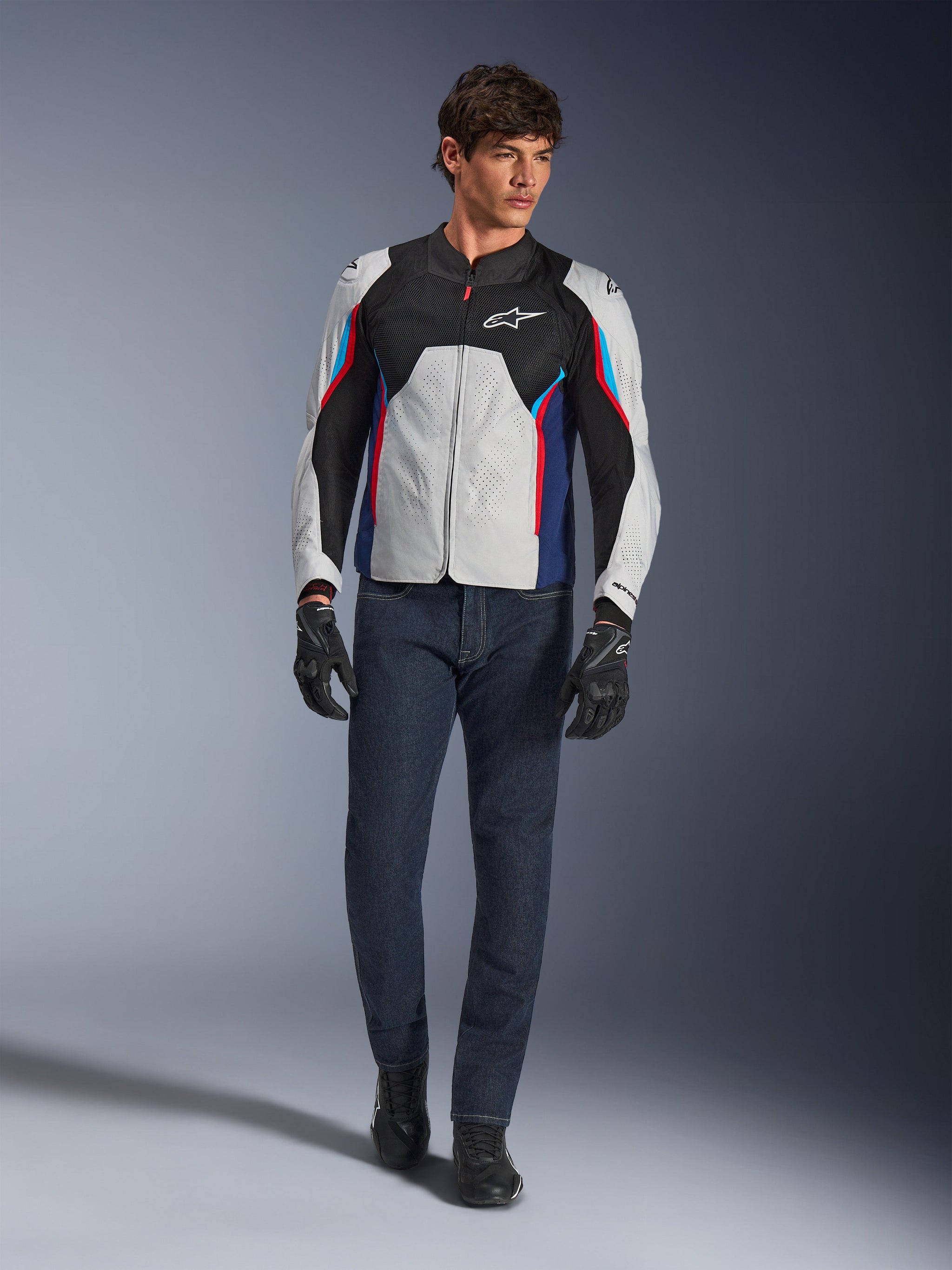 Modello uomo con giacca Alpinestars AST V3 Air in Silver Deep Blue Bright Red, caratterizzata da ventilazione in rete e pannelli traforati, abbinata a jeans da moto in denim scuro e guanti neri, equipaggiamento sportivo da moto per il clima caldo.