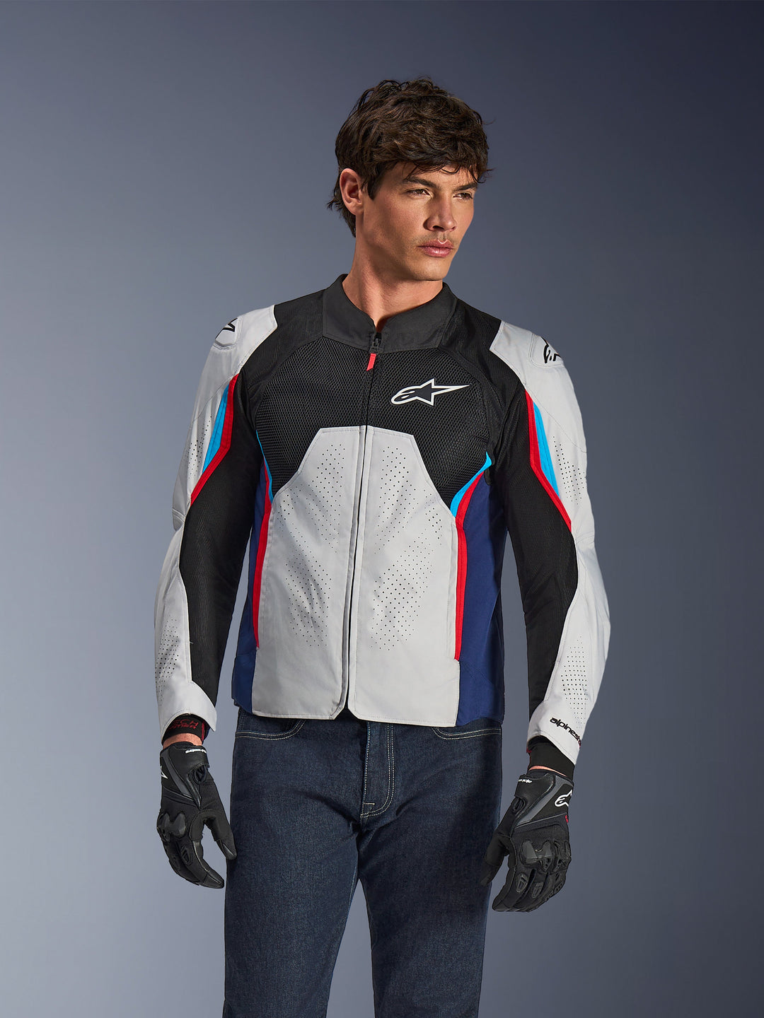 Giacca Alpinestars AST V3 Air in Silver Deep Blue Bright Red, indossata da un modello con jeans scuri e guanti neri, con pannelli argento traforati, ventilazione in mesh nero e accenti sportivi rossi e blu.