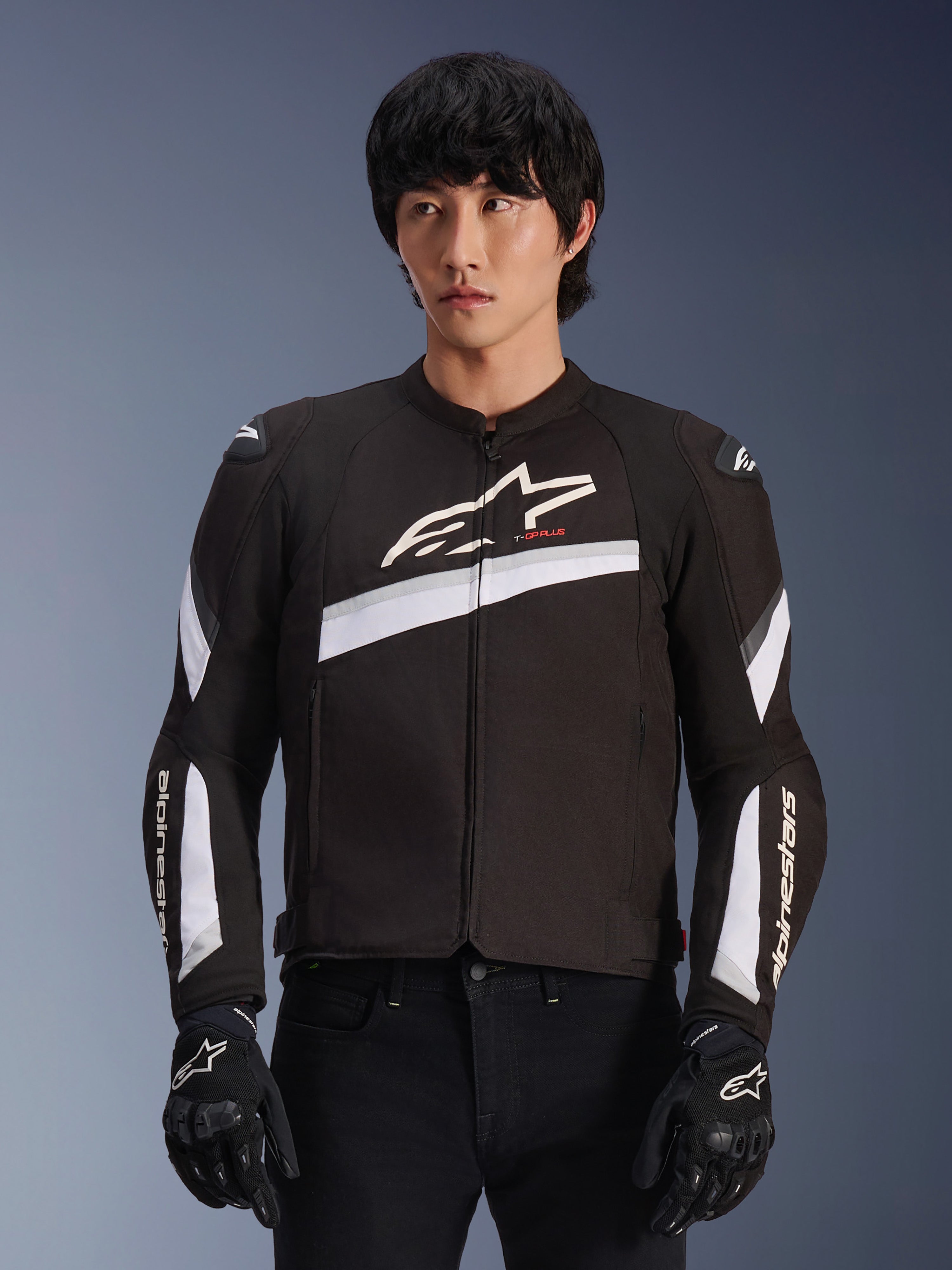 Giacca Alpinestars T-GP Plus R V4, nera e bianca, giacca da moto in tessuto, con accenti diagonali bianchi e loghi del marchio su petto e maniche, indossata da un modello con guanti da guida neri, design sport-fit aerodinamico per guida su strada