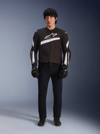 Giacca Alpinestars T-GP Plus R V4, nero e bianco, giacca da moto sportiva in tessuto, indossata da un modello con pantaloni e guanti neri, con vistoso logo Alpinestars bianco su petto e maniche, design elegante di ispirazione racing