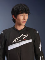 Giacca Alpinestars T-GP Plus R V4, nero e bianco, giacca da moto in tessuto indossata da un modello, con un grande logo a stella bianco sul petto, strisce diagonali bianche e protezioni per le spalle DFS Lite, abbigliamento da strada d'ispirazione sportiva