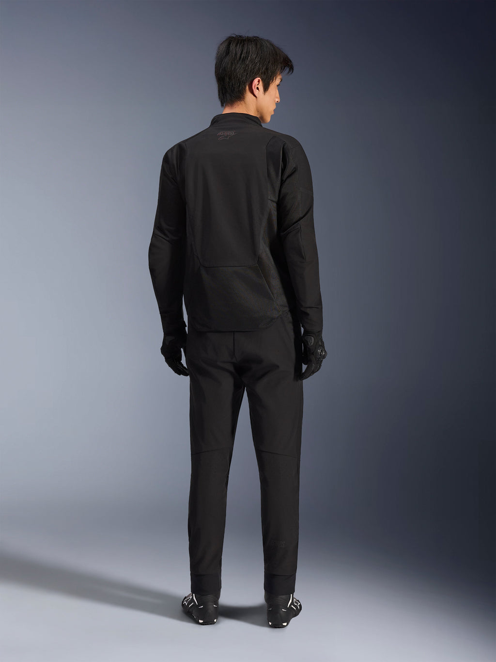 Giacca Alpinestars Aeroshell Airflow in Nero Antracite, vista posteriore di un modello maschile che indossa l'attrezzatura completa da moto inclusi pantaloni e guanti neri, con costruzione in softshell leggero e retro allungato per una maggiore copertura.