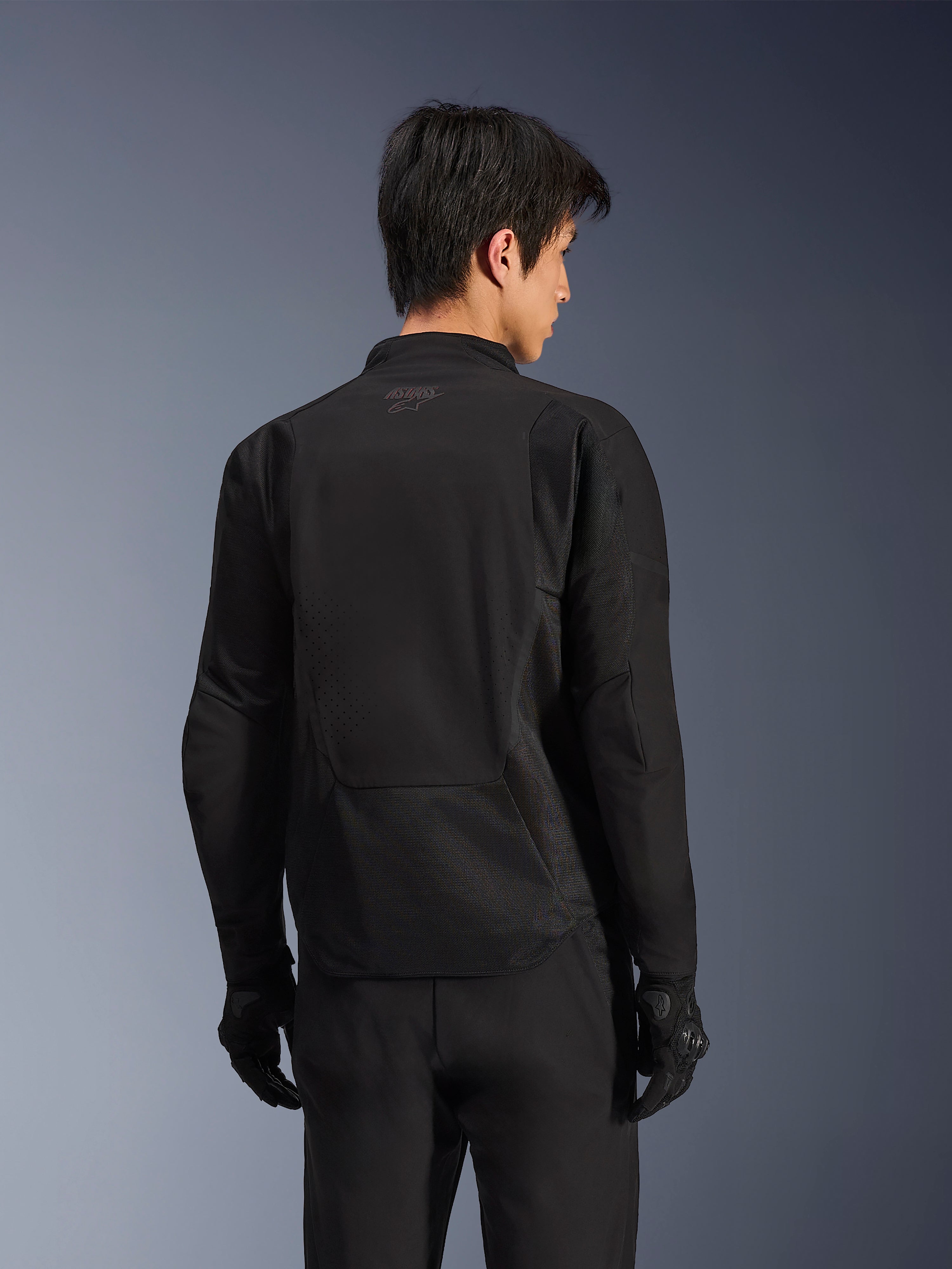 Giacca Alpinestars Aeroshell Airflow, Black Anthracite, vista posteriore di giacca da moto sportiva con zone di ventilazione perforate al laser e pannelli in mesh 2D, parte posteriore allungata per una maggiore copertura, indossata con guanti da guida neri