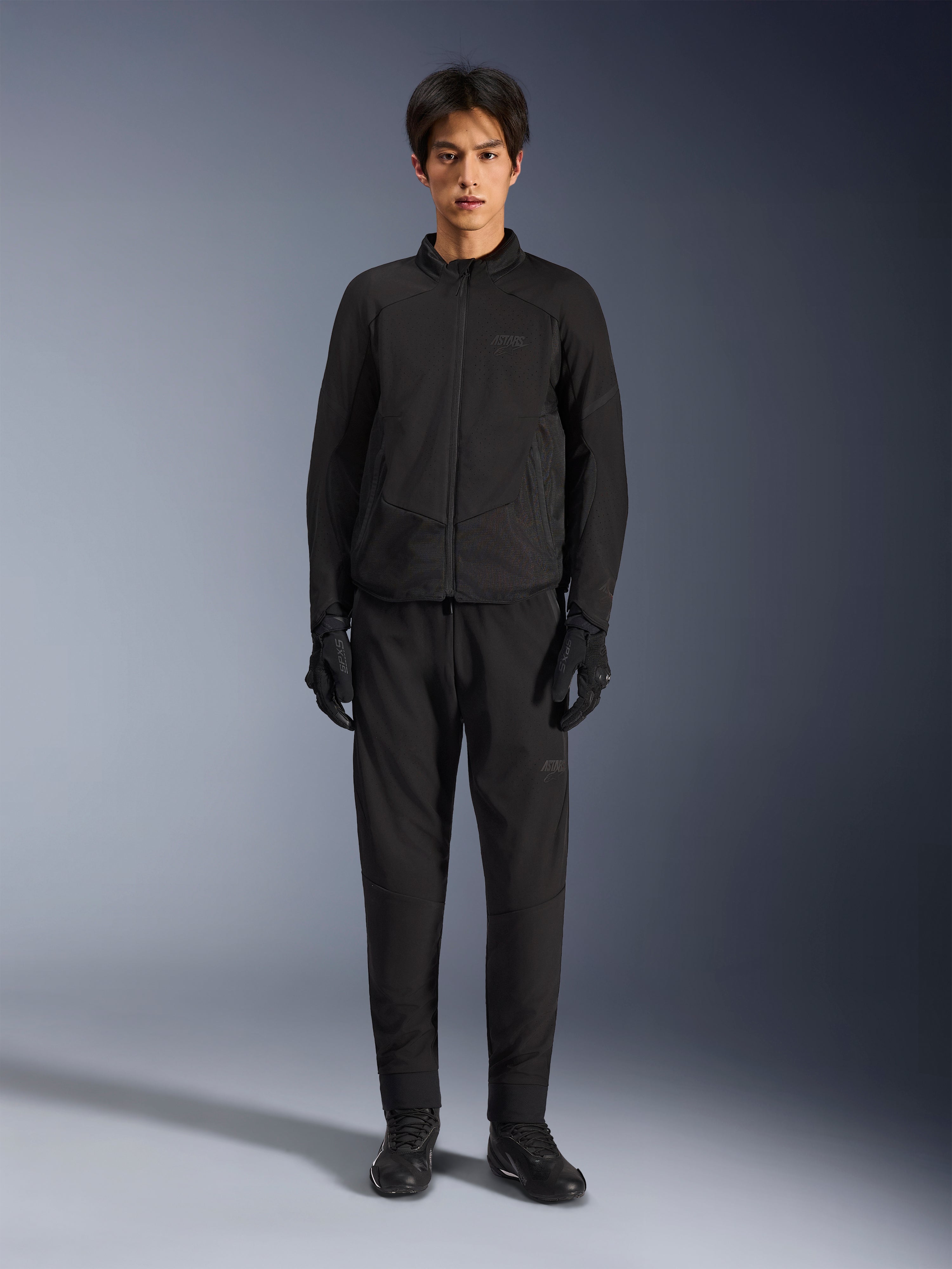 Giacca Alpinestars Aeroshell Airflow, giacca da moto nera, indossata da un modello con pantaloni e guanti neri coordinati, dotata di traforature laser sul petto e pannelli laterali in rete, vestibilità sportiva per prestazioni con climi caldi
