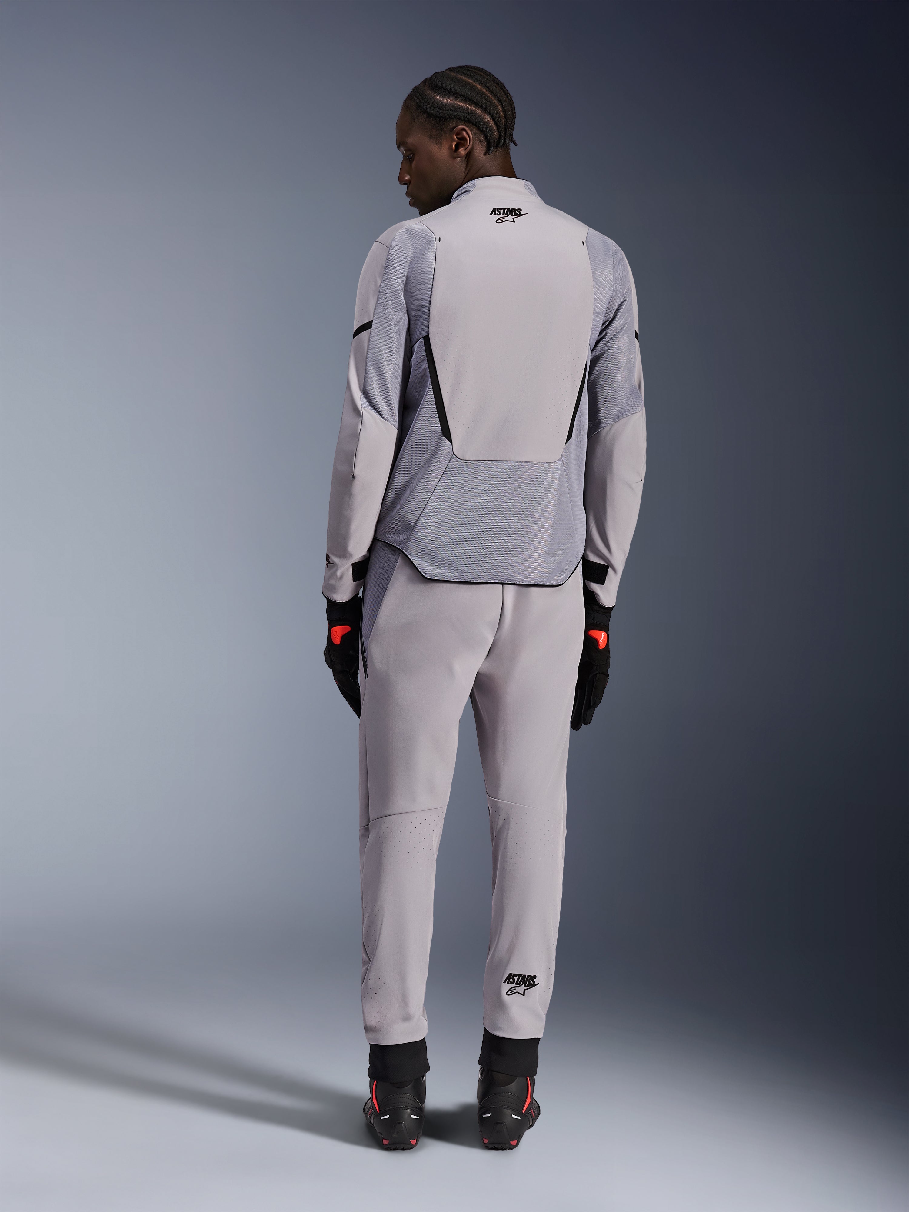 Giacca Alpinestars Aeroshell Airflow, Mid Gray Anthracite, vista posteriore su un modello, con pannelli in rete tecnica e zone di ventilazione perforate al laser, indossata con pantaloni grigi abbinati e attrezzatura da moto nera, design dal taglio sportivo per alte prestazioni.