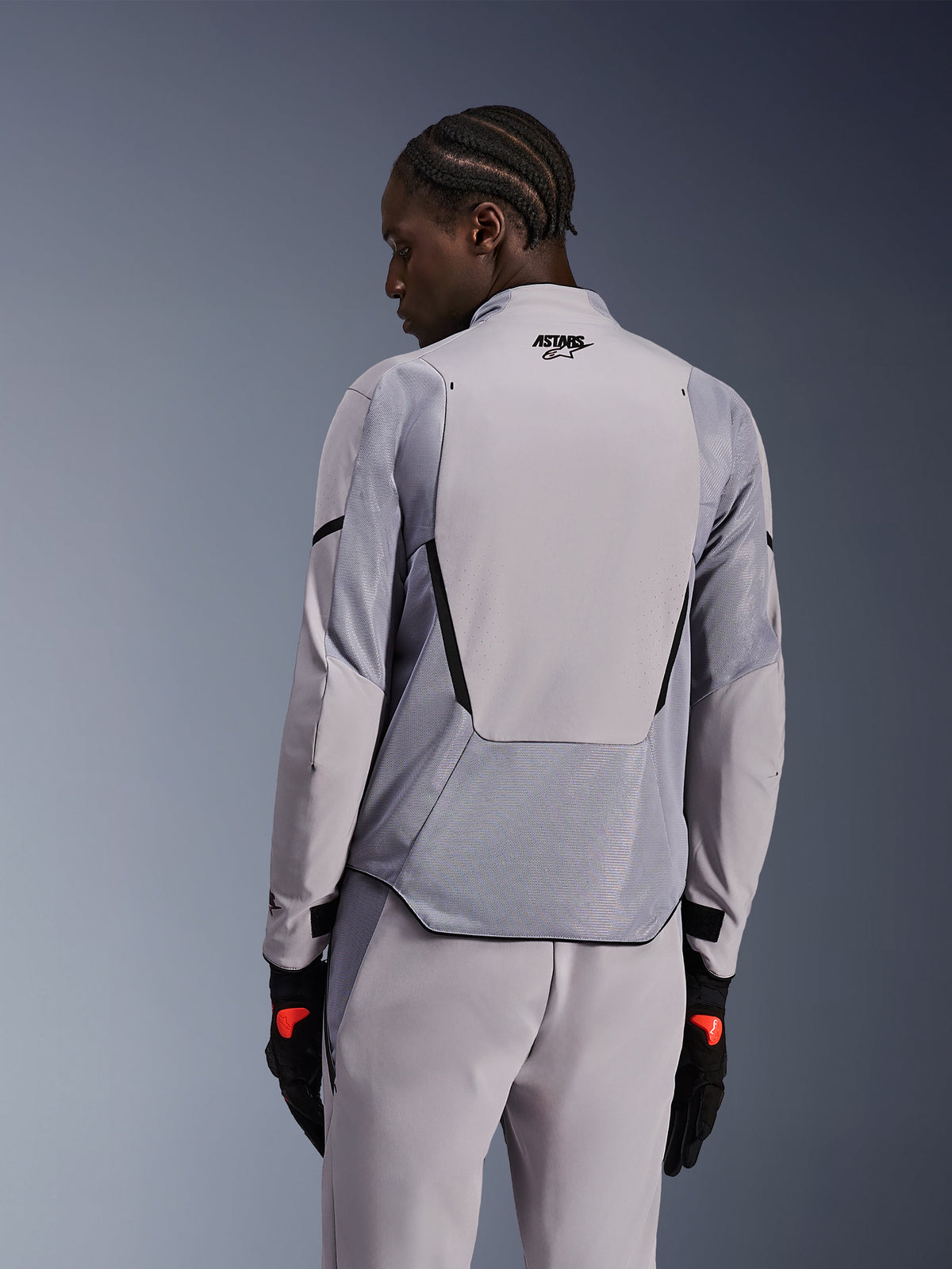 Giacca Alpinestars Aeroshell Airflow in grigio medio antracite, vista posteriore che mostra i pannelli in mesh traspirante e le perforazioni laser, costruzione tecnica in softshell elasticizzato con pannello posteriore allungato, indossata con pantaloni grigi coordinati e guanti neri per un look sportivo.