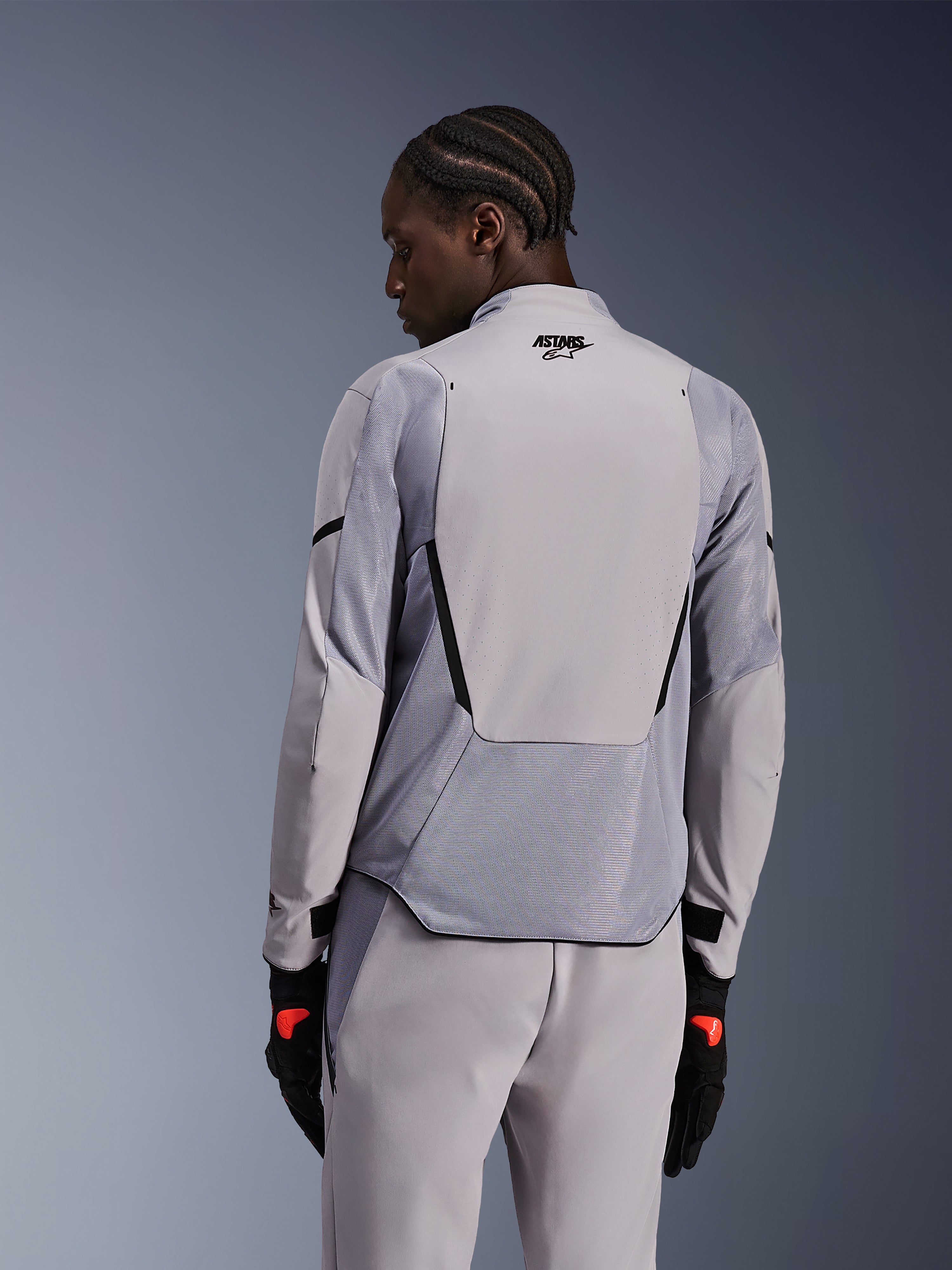 Giacca Alpinestars Aeroshell Airflow in grigio medio antracite, vista posteriore che mostra i pannelli in mesh traspirante e le perforazioni laser, costruzione tecnica in softshell elasticizzato con pannello posteriore allungato, indossata con pantaloni grigi coordinati e guanti neri per un look sportivo.