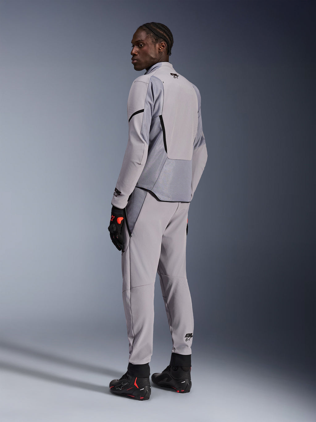 Giacca Alpinestars Aeroshell Airflow in Mid Gray Anthracite, indossata con pantaloni coordinati e guanti da corsa neri, vista posteriore che mostra i pannelli traforati al laser e la vestibilità sportiva atletica, abbigliamento professionale da moto
