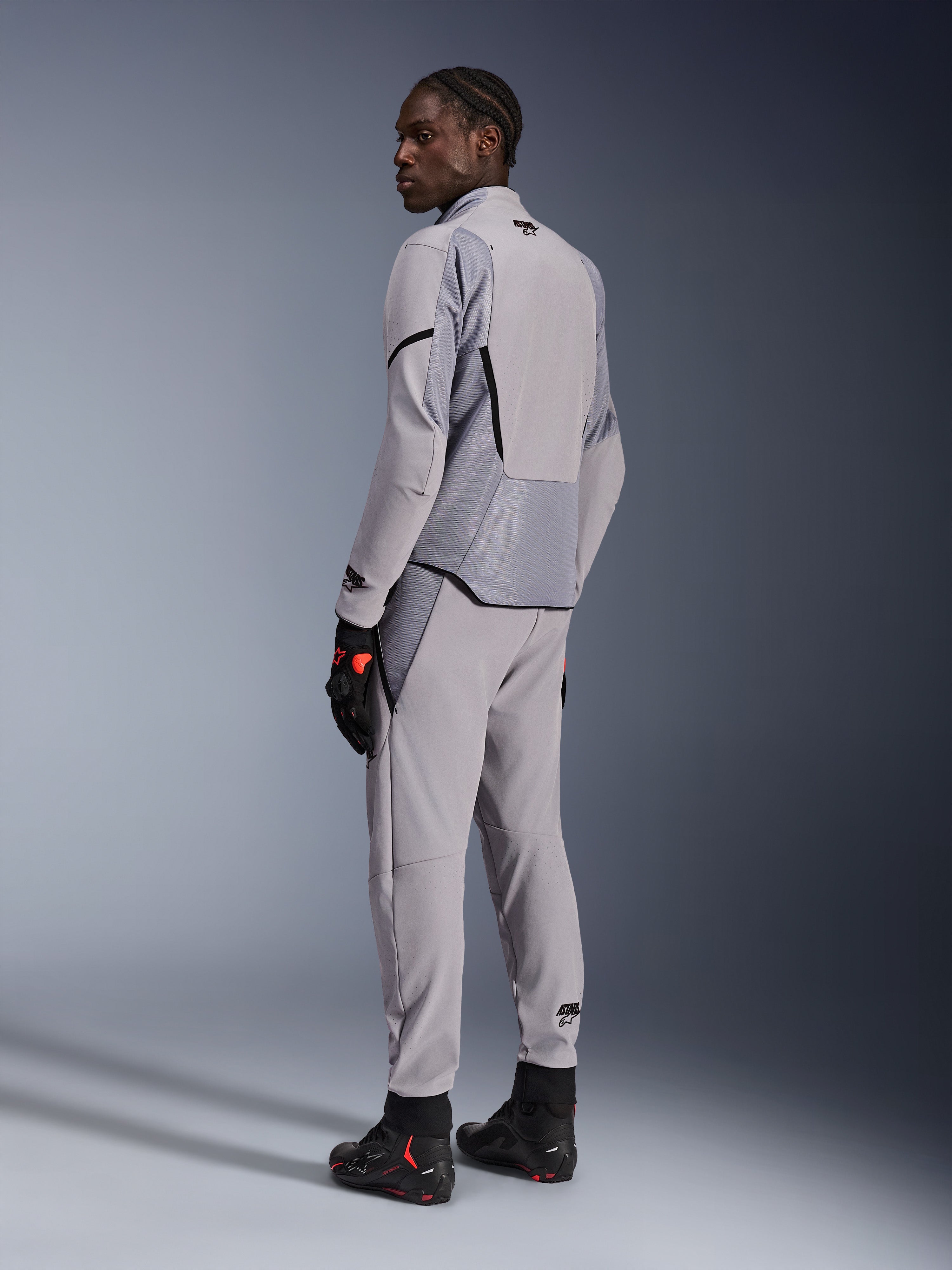 Giacca Alpinestars Aeroshell Airflow in Mid Gray Anthracite, indossata con pantaloni coordinati e guanti da corsa neri, vista posteriore che mostra i pannelli traforati al laser e la vestibilità sportiva atletica, abbigliamento professionale da moto