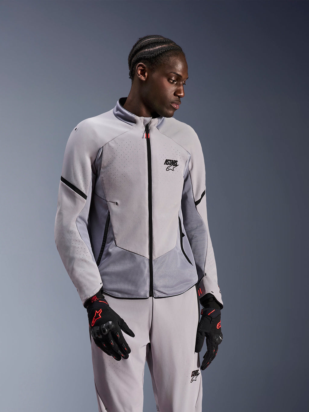 Giacca Alpinestars Aeroshell Airflow, grigio medio antracite, indossata da un modello con pantaloni in tessuto coordinati e guanti neri con accenti rossi, caratterizzata da zone perforate al laser e pannelli in rete per la ventilazione, design sportivo sport-fit per la guida in moto.