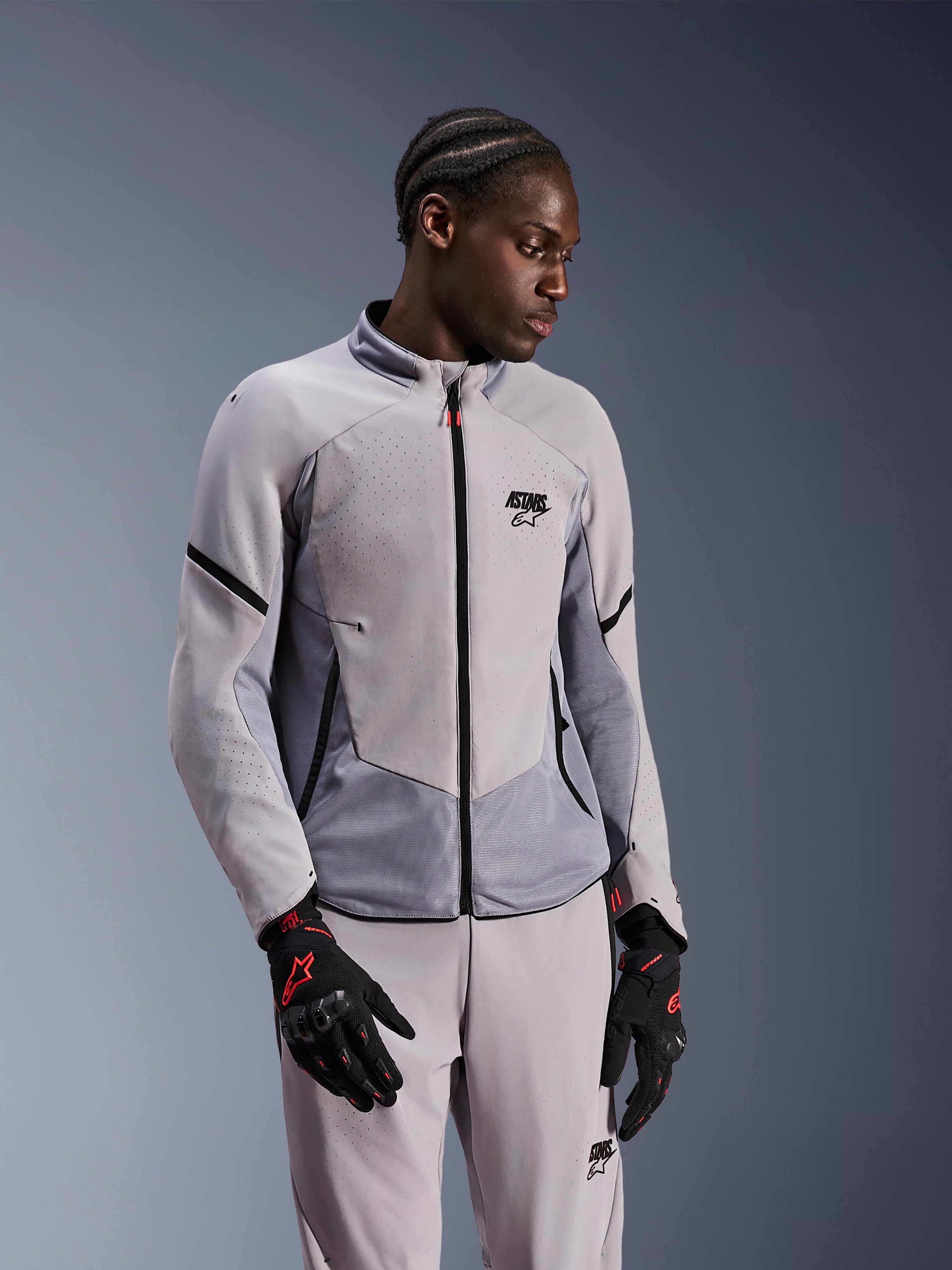Giacca Alpinestars Aeroshell Airflow, grigio medio antracite, indossata da un modello con pantaloni in tessuto coordinati e guanti neri con accenti rossi, caratterizzata da zone perforate al laser e pannelli in rete per la ventilazione, design sportivo sport-fit per la guida in moto.